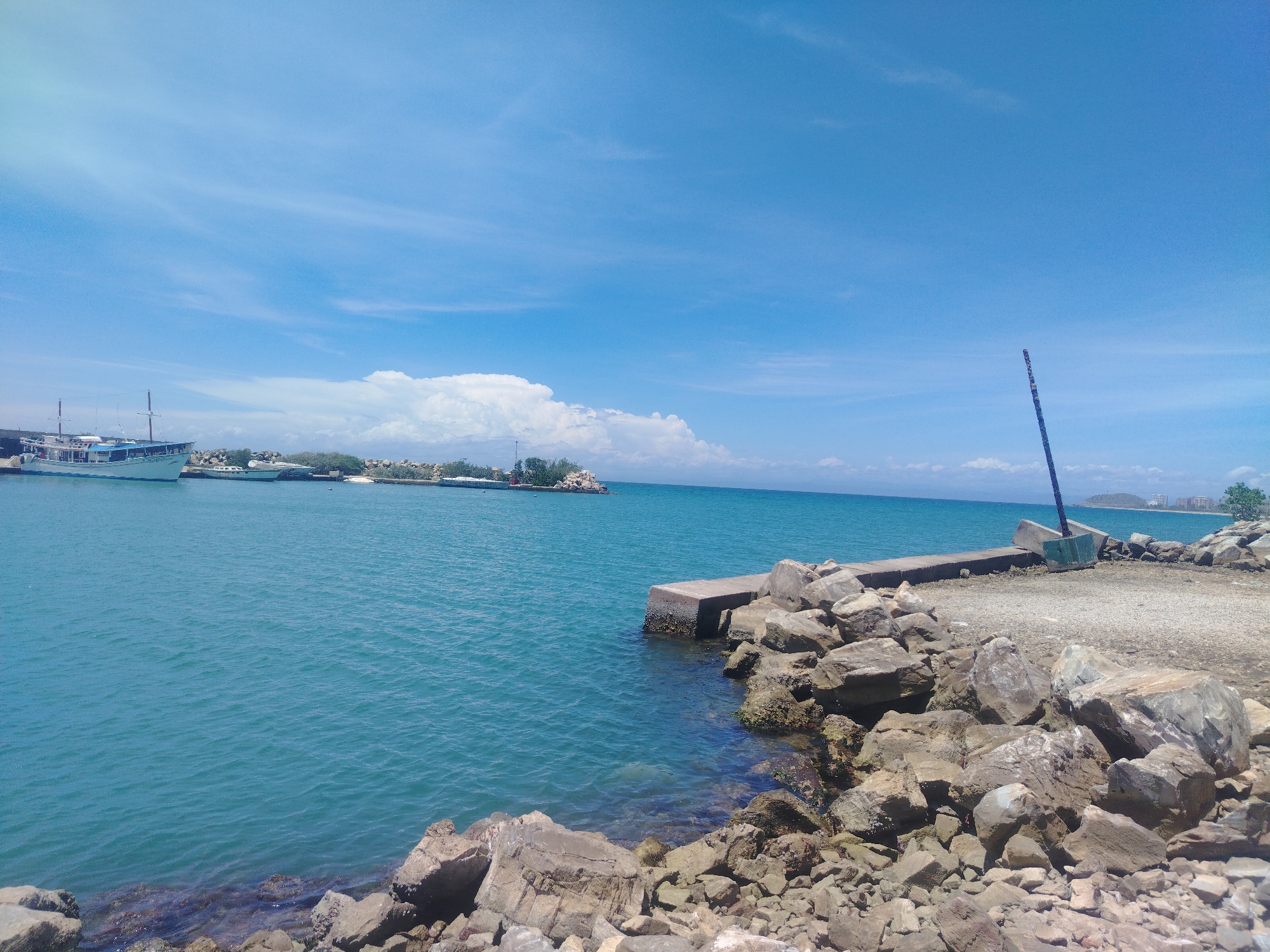 This media file depicts a bright, sunny day at a coastal location. The scene is dominated by a vast expanse of clear blue sky, with wispy white clouds scattered across it. Below the sky, the turquoise water of the ocean stretches out, appearing calm with gentle ripples on the surface.
On the left side of the frame, several boats are docked. One larger boat, with a white hull and multiple decks, is prominently visible. Behind it, other vessels can be seen, suggesting a harbor or marina. The water in this area is a vibrant blue-green.
The foreground of the image is occupied by a rocky shoreline. Large, irregular stones and pebbles are piled along the edge of the water, creating a rugged and natural barrier. A concrete structure, possibly a pier or part of a breakwater, juts out into the water, also surrounded by rocks. A dark, slender pole, possibly a marker or part of a structure, stands upright on the concrete in the right portion of the image.
In the distance, across the water, a line of trees and some buildings can be seen along the horizon, hinting at a populated area or a town. The overall impression is one of a peaceful and picturesque seaside setting on a clear day. The sunlight suggests it is midday. There are no people visible in the image. No text is discernible.
