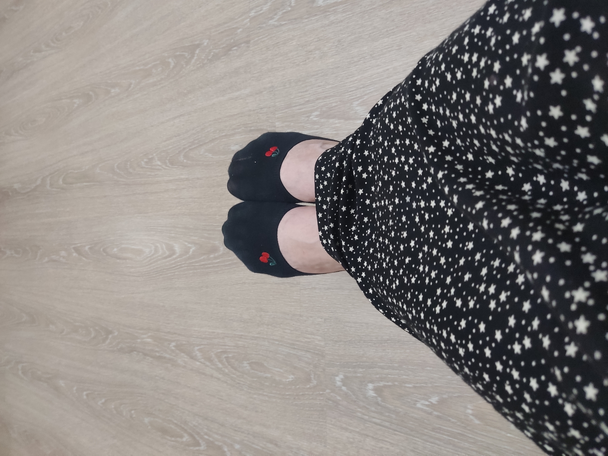 The image shows a pair of feet wearing black no-show socks with embroidered red cherries on them, standing on a light-colored wooden floor. The lower part of a black skirt with a white star pattern is visible in the foreground. The scene is indoors, and there is no indication of the city of Kurchaloy, Russia.