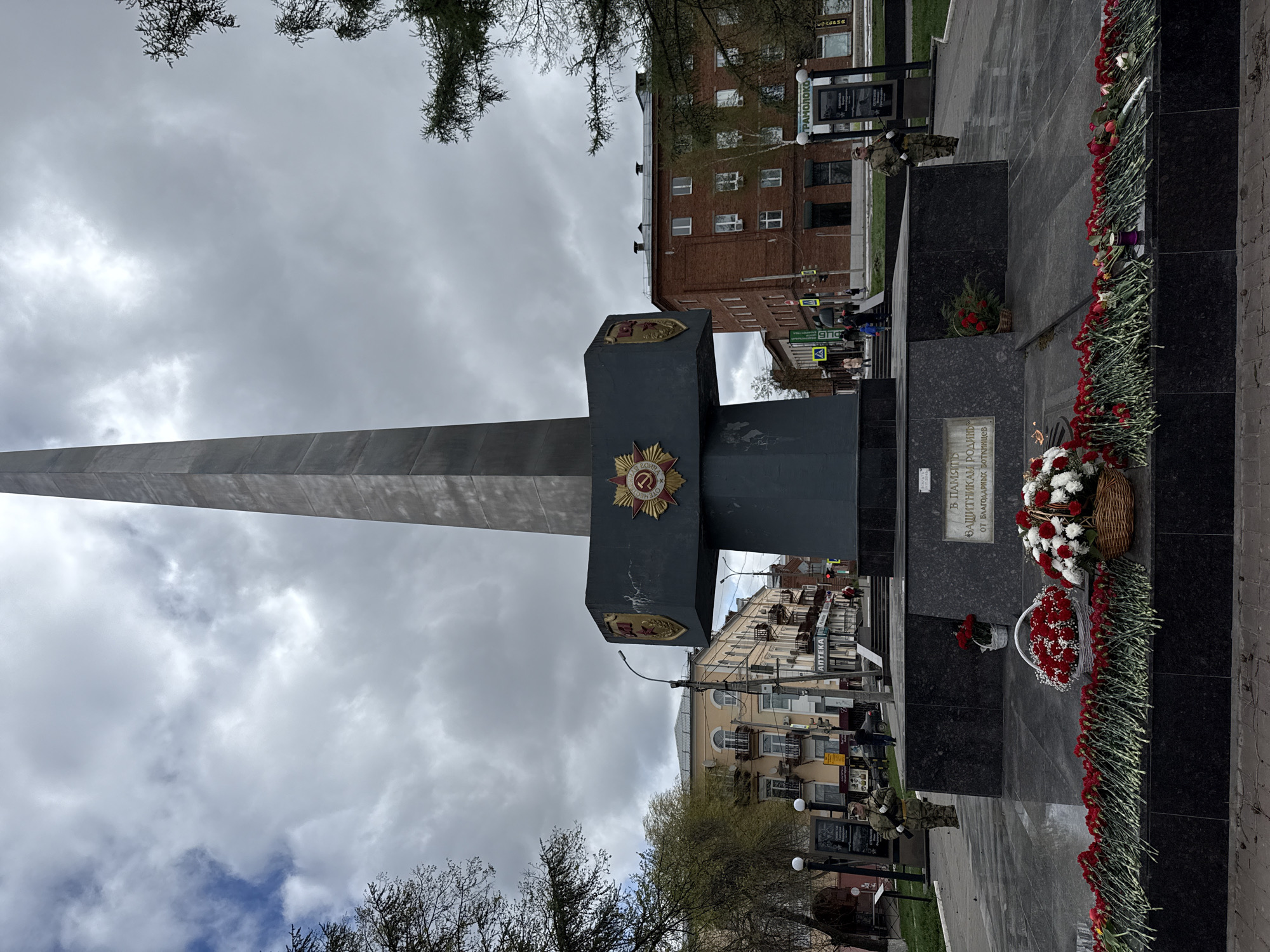 The image depicts the Victory Monument in Votkinsk, Russia, a tall, grey obelisk with a dark, cubic structure intersecting it horizontally, adorned with Soviet-era emblems. The monument stands in a paved square, with bouquets and baskets of red and white flowers placed at its base. A plaque with Cyrillic text is visible on the structure. Buildings line the background, including a brick building with multiple windows and a lighter-colored building with signs in Russian. Trees with sparse foliage are in the foreground, and an overcast sky with grey clouds is above. A person in military fatigues is standing near the monument. Several streetlights are visible.