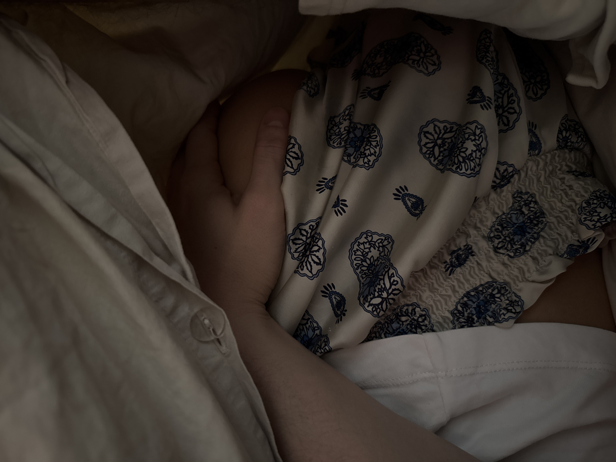 A close-up shot taken indoors shows a person's hand gently holding a bare shoulder. The person is wearing a white and blue patterned top with an elasticized section. Part of a light-colored button-down shirt is visible in the lower left. The location appears to be in an unknown city, province, and state.