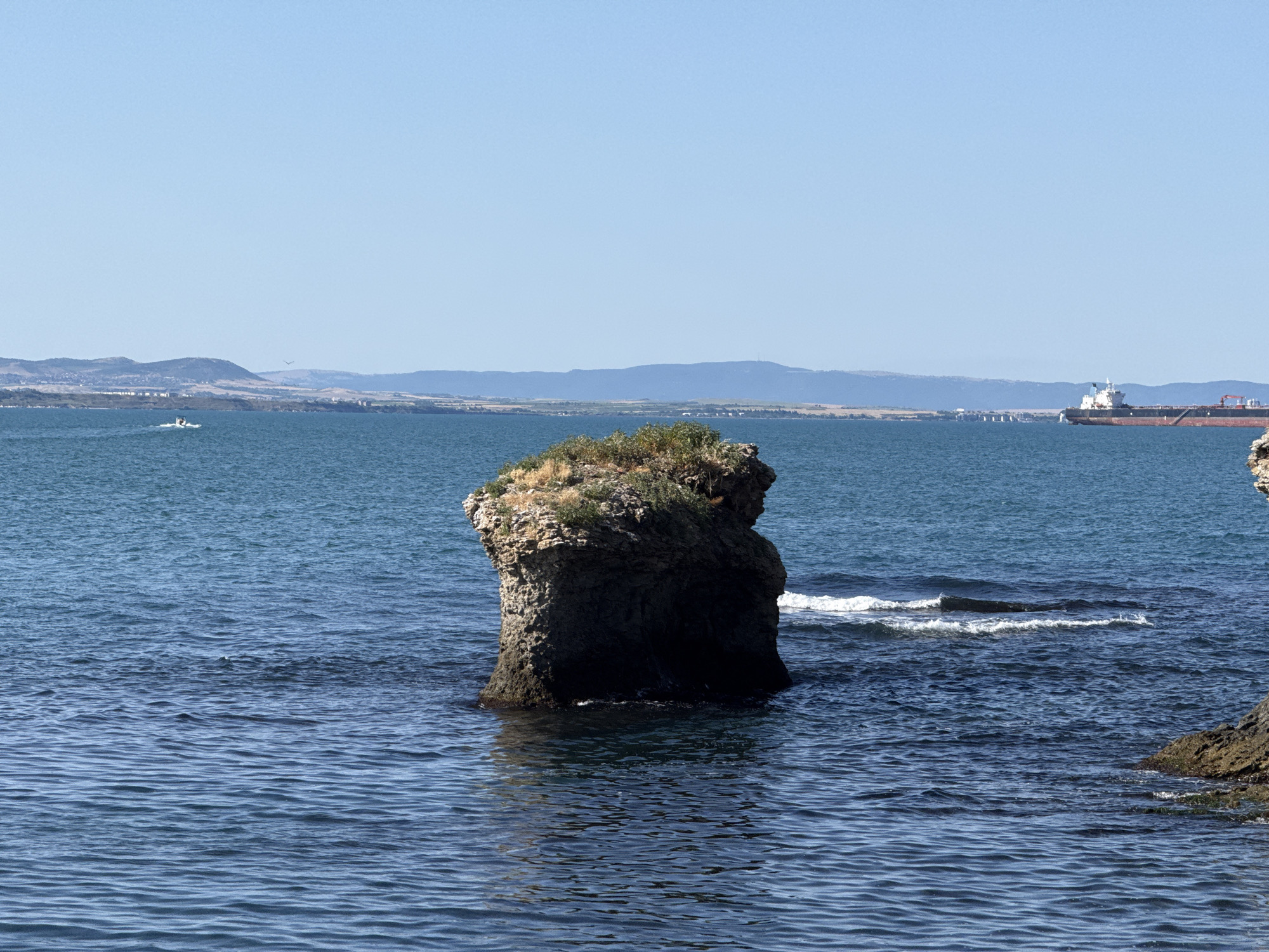 The image depicts a rocky outcrop in the sea, likely off the coast of Burgas, Bulgaria, given the context of the prompt and visual cues of the landscape and the presence of a large cargo ship in the distance. The sea is a deep blue with moderate ripples. The rock formation in the foreground is substantial, with a rugged texture and vegetation growing on its crest. The water around the rock shows some small waves and white foam.
In the background, a distant landmass is visible, characterized by rolling hills and scattered buildings suggesting a coastal town or city. The sky is clear and a pale blue. Further out at sea, a large cargo ship is visible, its white superstructure and dark hull contrasting with the blue water. A small white boat is also visible in the middle distance, creating a wake. The overall scene is one of a serene coastal environment under a bright sky.