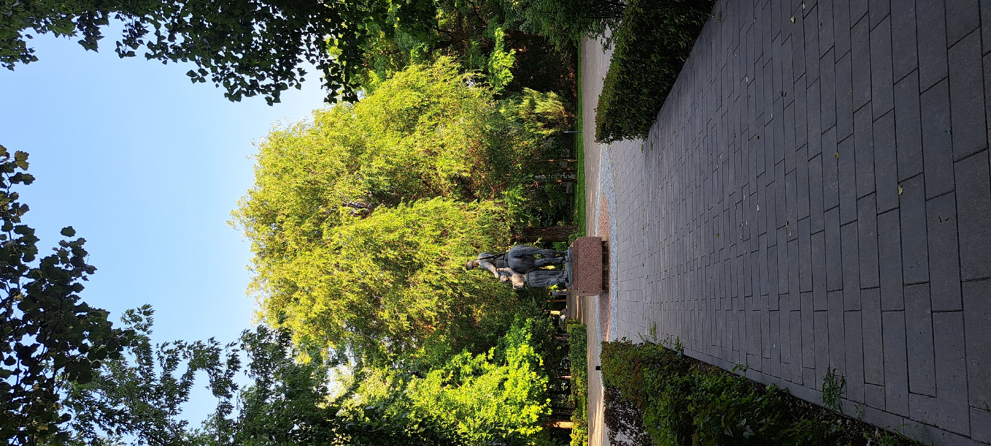 A bronze equestrian statue is positioned on a stone pedestal in a park. The statue depicts a man in military attire on horseback, with a woman standing next to the horse. The scene is surrounded by lush green trees and manicured hedges, with a paved walkway leading towards the statue. The sky is clear and blue. The overall impression is one of a peaceful, well-maintained public space.