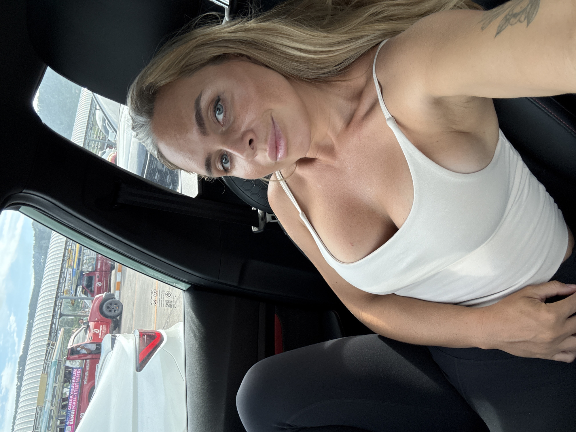 A blonde woman with blue eyes and fair skin is sitting in the passenger seat of a car, looking at the camera. She is wearing a white tank top and black leggings. Her left arm is resting on her lap, and her right arm is extended towards the camera. The background shows a street scene with several vehicles, including a red truck with text that reads "CHECK YOUR HOLES! SAMUI PCR14 1 TEST 14 DAYS". The scene appears to be a checkpoint or a testing facility. The car is parked, and the sun is shining. The location is identified as the city of Ko Samui District, Thailand.