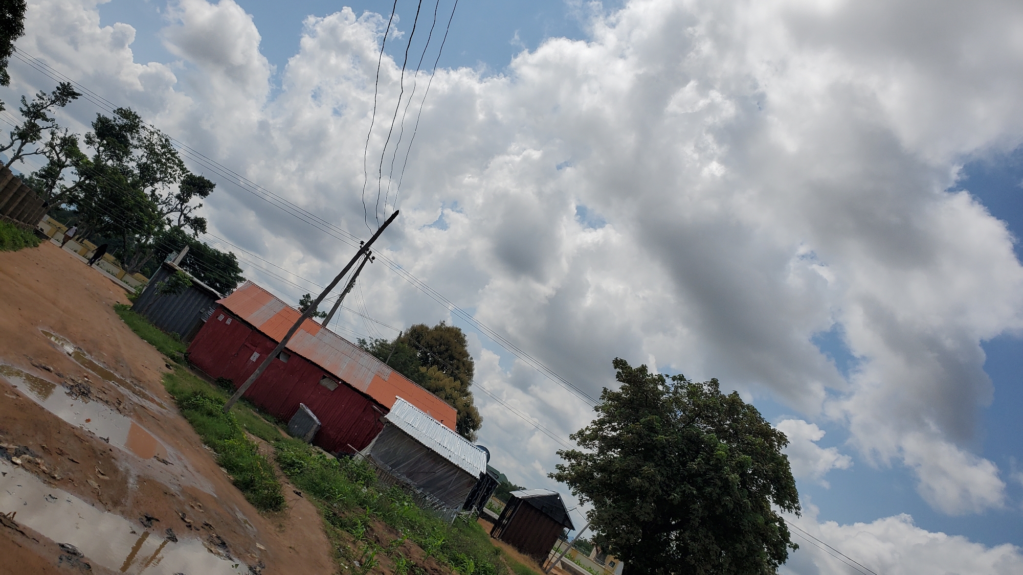 The image depicts an outdoor scene with a predominantly cloudy sky. In the foreground, a muddy and uneven dirt road is visible, with several large puddles reflecting the sky. To the right of the road, there is a red building with a corrugated metal roof. Behind this building, a smaller, dark structure, possibly a shed or kiosk, can be seen. Vegetation, including grass and trees, lines the area around the buildings. Utility poles with multiple wires are prominent in the middle ground. The background shows more trees and a distant building. The perspective of the image is tilted, giving the impression of a slight upward angle. This scene is likely located in the city of Jalingo, Nigeria, given the common appearance of such rural or semi-urban settings in the region, characterized by unpaved roads, simple structures, and abundant vegetation under a tropical sky.