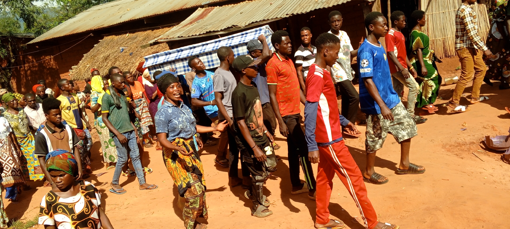 The image shows a procession of people in Ngalula, Democratic Republic of the Congo. Many people are walking on a red dirt road. A group of men are carrying a mattress above their heads, while others walk alongside. The individuals are wearing a variety of clothing, some in traditional African attire and others in modern casual wear. Some people appear to be talking and smiling. In the background, there are traditional buildings made of clay or mud bricks with straw roofs. The overall environment suggests a rural setting within the city of Ngalula.