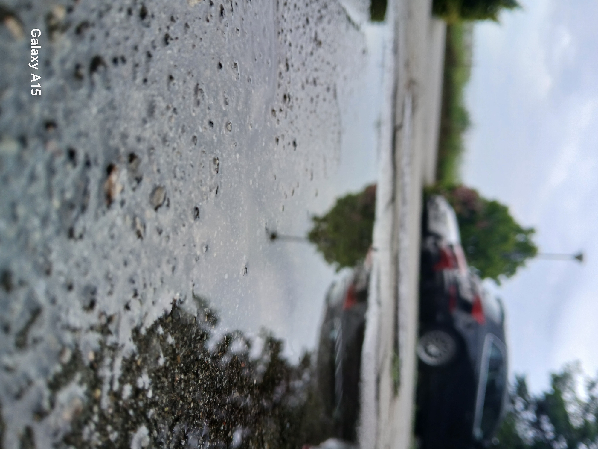 An extreme low-angle close-up shot focuses on a puddle of water on asphalt, reflecting the overcast sky and surrounding environment. In the background, out of focus, are parked cars and greenery, suggesting an outdoor urban setting. The watermark "Galaxy A15" is visible in the lower left corner.
