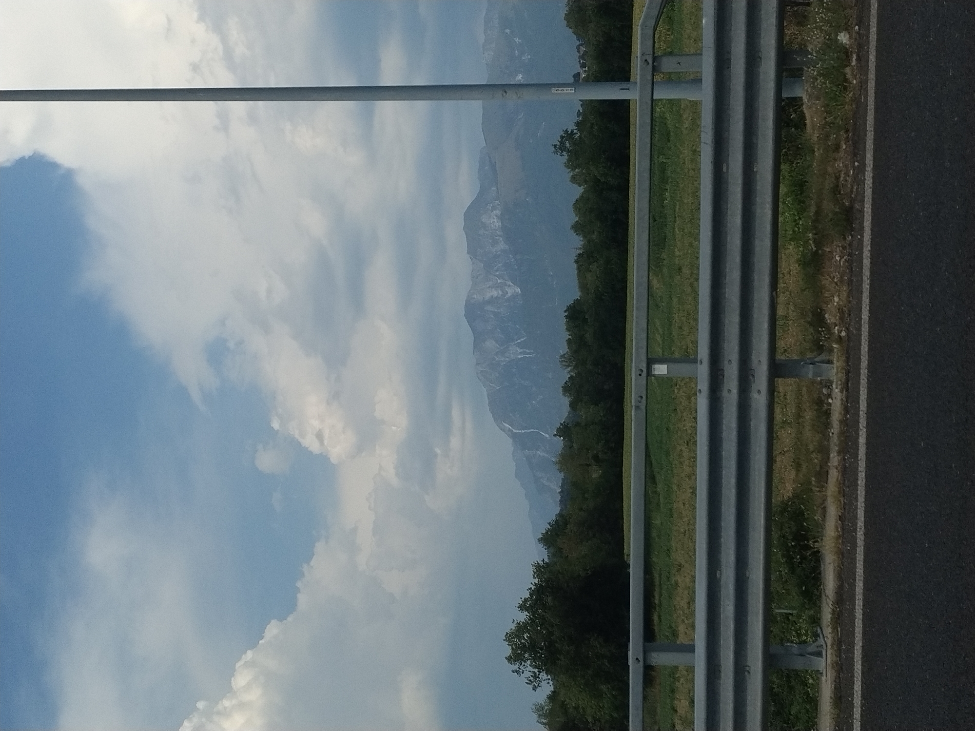 The image depicts a roadside scene with a guardrail in the foreground, an asphalt road, and a grassy embankment. Beyond the guardrail, a dense line of trees forms a natural barrier, and behind the trees, a majestic mountain range with snow-capped peaks dominates the horizon. The sky above is a mixture of blue and white clouds, with some darker, more ominous clouds suggesting a potential storm. A tall, grey metal pole, likely a lamppost, stands to the right of the frame, bisecting the view of the mountains. The overall impression is one of a natural landscape viewed from a man-made structure, possibly a highway. No people are visible in the image. The location in Mels, Italy is not explicitly indicated by any visual cues in the image itself.