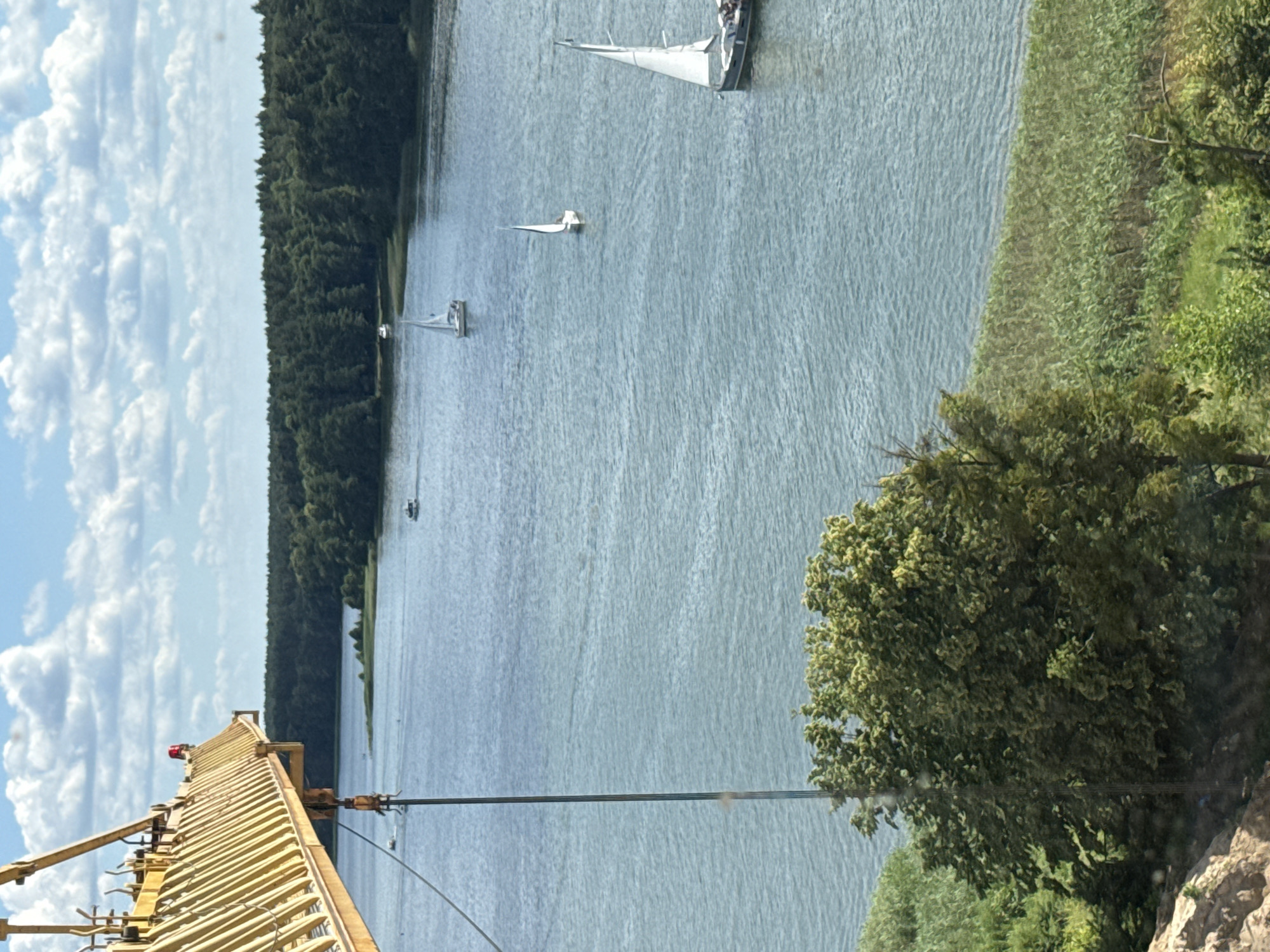 This media file depicts a wide shot of a body of water, likely a lake or bay, with a dense forest lining one side. Several sailboats are visible on the water, suggesting recreational sailing. In the foreground, a yellow crane structure extends from the left side of the frame, with a red light visible on its top. A body of water and a tree with green foliage are also visible to the right. The sky is partly cloudy. The overall scene suggests a pleasant, possibly sunny day. The image appears to be taken from an elevated perspective, possibly from the crane itself or a nearby elevated structure. No visible text is present in the image. The location context of Mikołajki, Poland, known for its lakes and sailing activities, aligns with the visual elements.