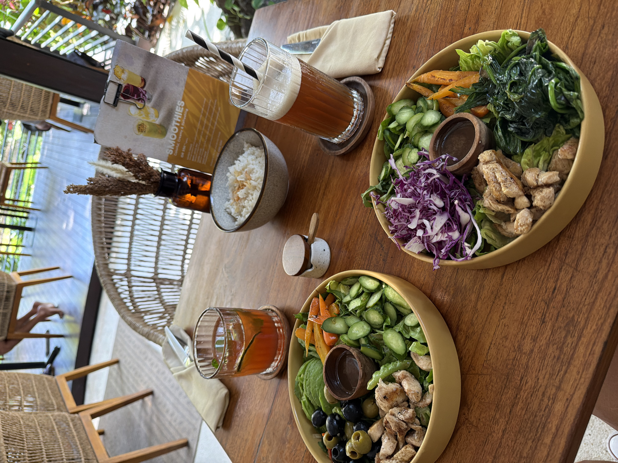 Two healthy meal bowls are displayed on a wooden table at a restaurant in Ubud District, Indonesia. The bowl on the left contains chicken, olives, avocado, cucumber, carrots, and greens. The bowl on the right holds chicken, red cabbage, cucumber, carrots, spinach, and greens. A small wooden cup with a dark sauce sits in the center of each bowl. In the background, a glass of iced tea and a glass of a red-colored beverage are visible. A menu with smoothie options is propped up behind the iced tea. Two wicker chairs are partially visible in the foreground and background. The overall ambiance suggests an outdoor dining setting with natural lighting and lush greenery.