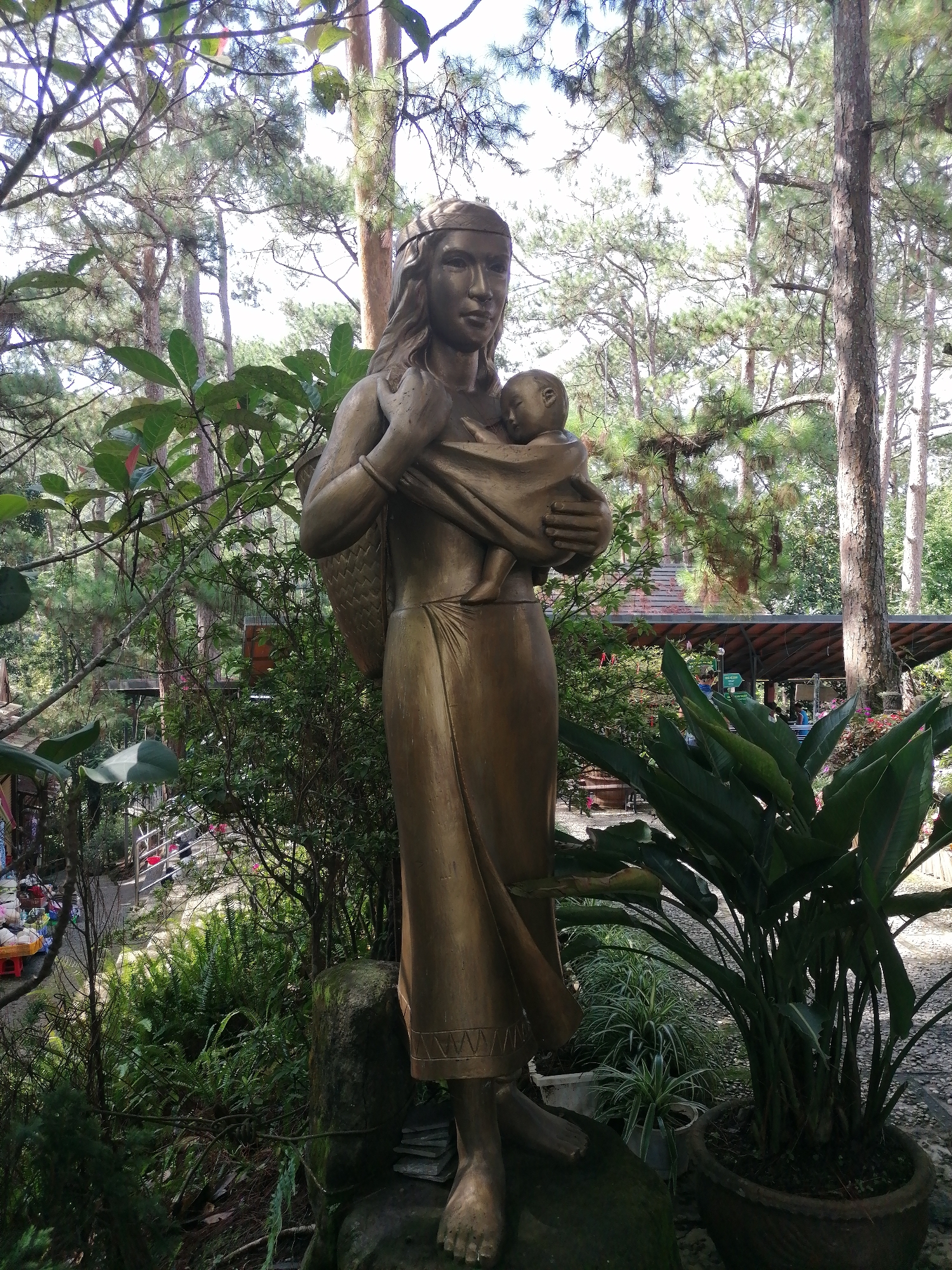 This is a photograph of a bronze statue of a woman holding a baby, set in an outdoor environment with trees and foliage. The statue is the primary subject, positioned centrally and facing the viewer. The woman wears a headdress and appears to be dressed in traditional attire. She is holding a baby wrapped in a cloth, cradled against her chest. Behind and around the statue are tall pine trees, characteristic of the cool climate in Da Lat, Vietnam. The ground is covered with a mixture of gravel and vegetation, including ferns and potted plants. In the background, there's a glimpse of a covered structure, possibly a pavilion or stall, with some people in the distance. The overall impression is a serene, natural setting, likely a park or a tourist attraction in Da Lat, a city known for its pine forests and cool temperatures, similar to the French Alps.