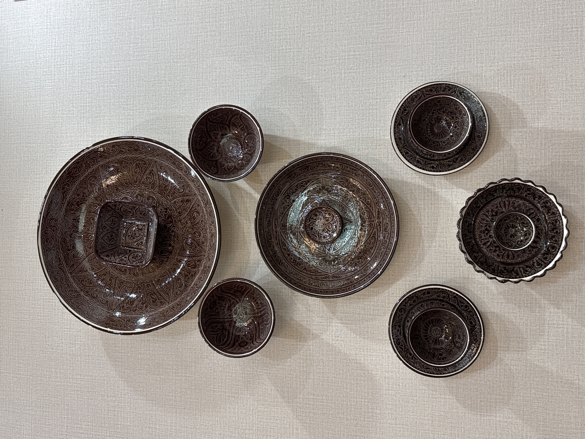 The image shows a collection of seven brown ceramic dishes with intricate patterns displayed on a light beige textured wall. The arrangement includes a large bowl with a square centerpiece, a medium-sized plate, two small bowls, and three small plates, one of which has a scalloped edge. The dishes appear to be of Uzbekistani design, likely from Samarkand City, Uzbekistan, known for its pottery traditions. There are no people visible in the image. The lighting suggests an indoor setting, possibly a store or a private collection.