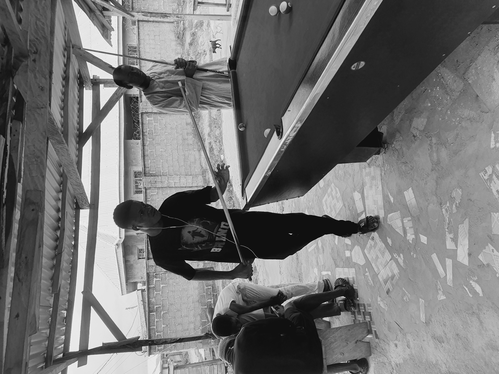 The black and white image captures an outdoor billiards game in Lokoja, Nigeria, under a makeshift roof. In the foreground, a young man in a t-shirt and shorts is poised to strike a billiard ball with a cue stick, his eyes focused on the game. To his right, another man, dressed in traditional attire, holds his own cue, observing. In the background, two individuals are seated on a wooden bench, seemingly engaged in conversation or observing the match. The ground is a mosaic of broken tiles and concrete, a common sight in many outdoor spaces in Lokoja. The surrounding environment features a brick wall, barbed wire, and the roofs of houses, indicating an urban setting. Two dogs are visible in the mid-ground, adding a touch of local life to the scene. The overall atmosphere suggests a casual gathering and a moment of leisure in the heart of Lokoja.