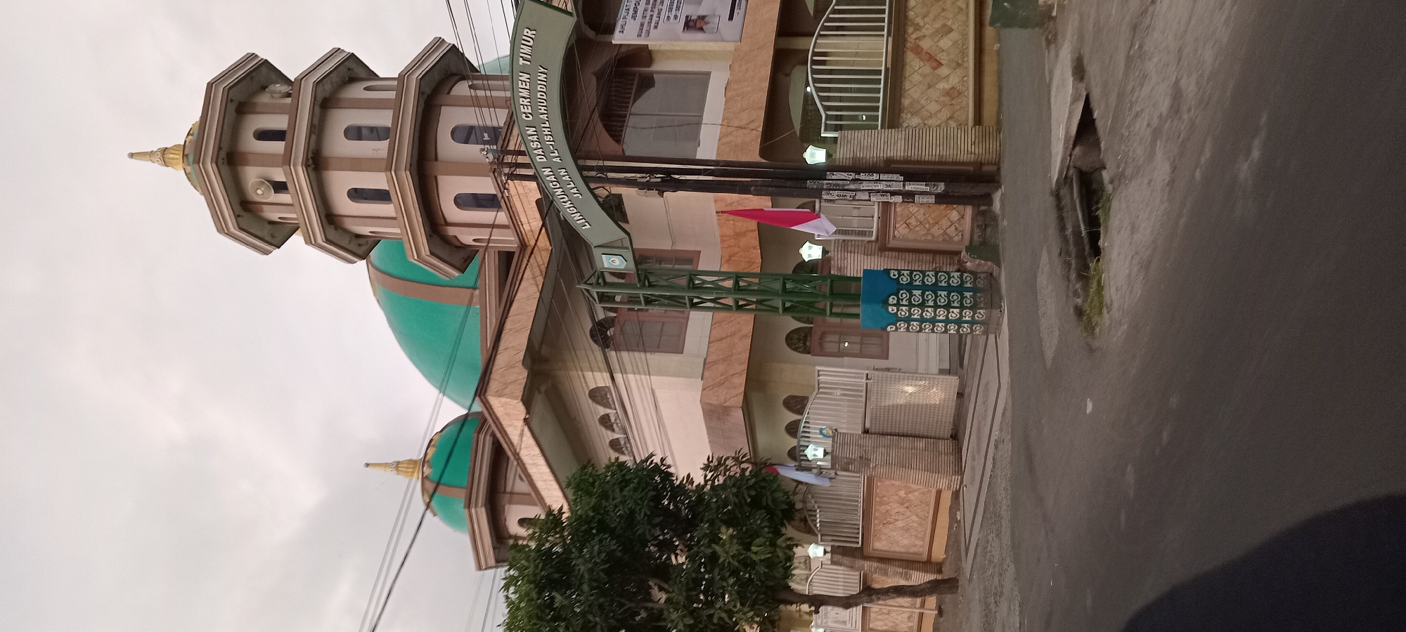 The image shows a mosque in the city of Mataram, Indonesia. The mosque has a prominent green dome and a tall, multi-tiered minaret. An archway above the entrance reads "LINGKUNGAN DASAN CERMEN TIMUR JALAN AL-ISLAHUDDINNY," indicating the mosque is located in the Dasan Cermen Timur area on Jalan Al-Islahuddiny. A red and white Indonesian flag is flying from a pole. The mosque is surrounded by a decorative wall and fence. In the foreground, a paved road is visible, with a drain in the lower right corner. The sky is overcast.