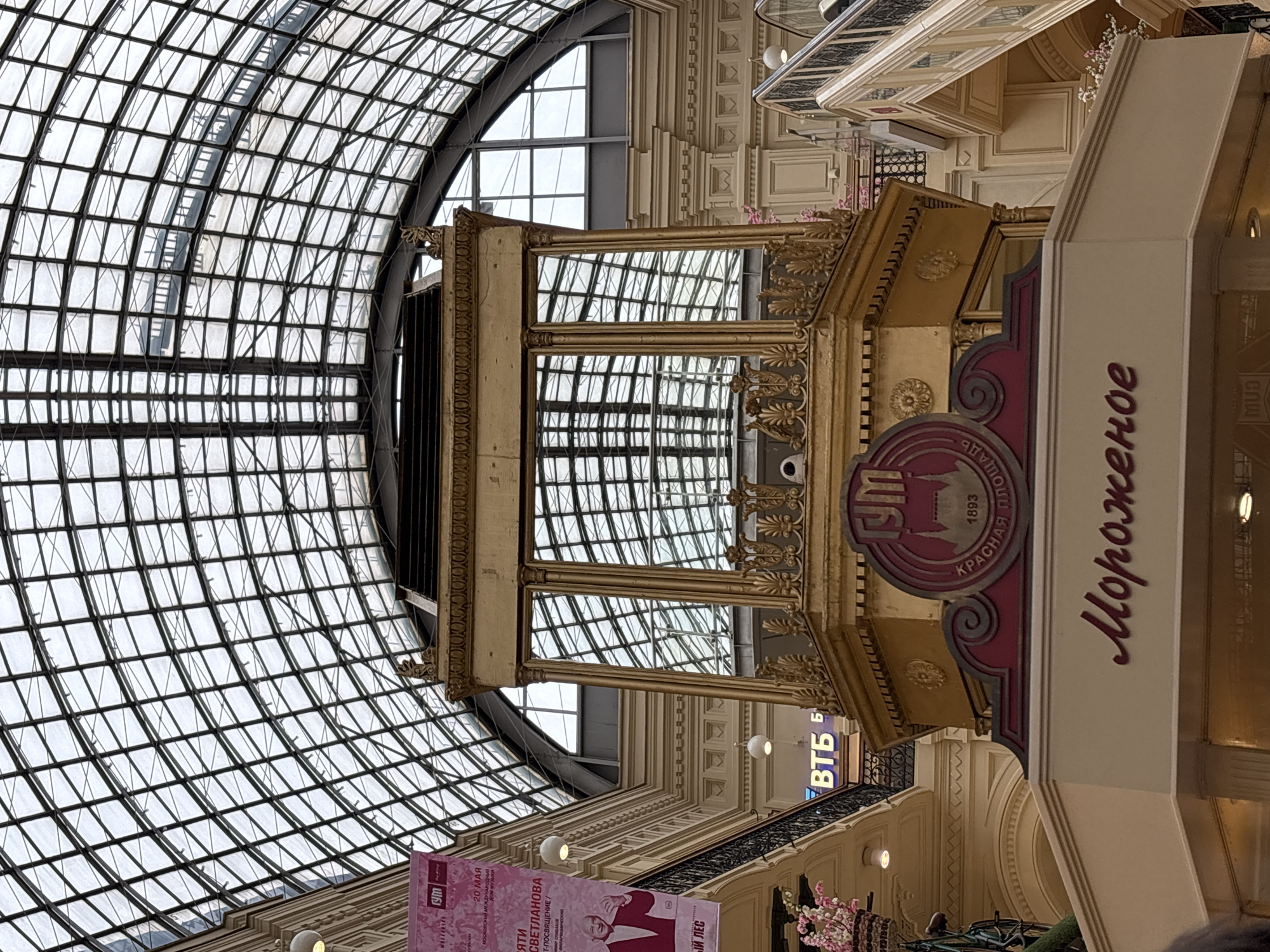 The image shows an indoor scene with a large, arched glass ceiling. Beneath the ceiling, there is a large, ornate, gold-colored structure with mirror panels that reflect the glass ceiling. To the right of the golden structure is a sign with the words "Мороженое" vertically written in Russian, meaning "Ice Cream" in English. The sign also includes the Russian abbreviation for "GUM" or "ГУМ" (state department store), the year "1893", and the words "КРАСНАЯ ПЛОЩАДЬ" which means "Red Square". Further in the background and below the sign is a "VTB" bank logo. The scene takes place in GUM, located in Moscow, Russia.