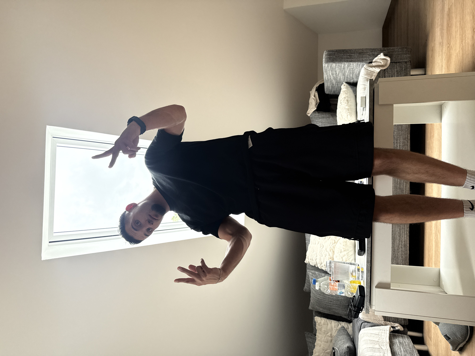 A man is standing in a room with a slanted ceiling and a large skylight. He is wearing a black t-shirt and black shorts. His right arm is raised, with his index and middle fingers extended, forming a peace sign. His left arm is also raised, with his index and middle fingers extended, and his thumb tucked in, forming a different gesture. He is standing on top of a white table that is in front of a gray sofa with several pillows. On the table, there is a water bottle, a glass, and some electronic devices. The floor is wooden. The image was taken in Garmisch-Partenkirchen, Germany.