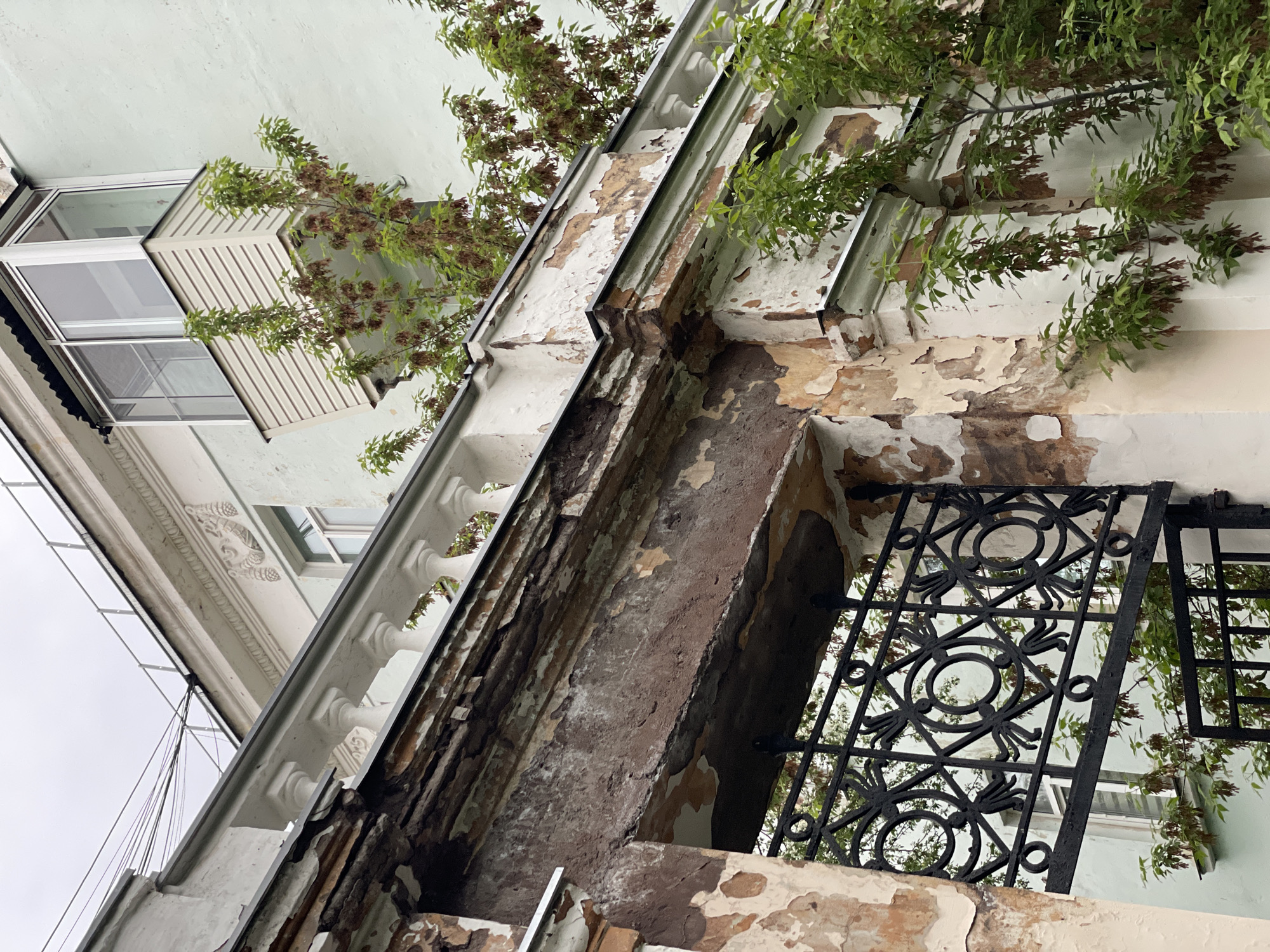 The image depicts a deteriorating exterior of a building, showing flaking paint, exposed brick, and some foliage. A wrought-iron balcony railing is also visible. In the background, another building with a window and some decorative molding can be seen. The sky is overcast. There is no evidence to support a connection to Zheleznogorsk, Russia.