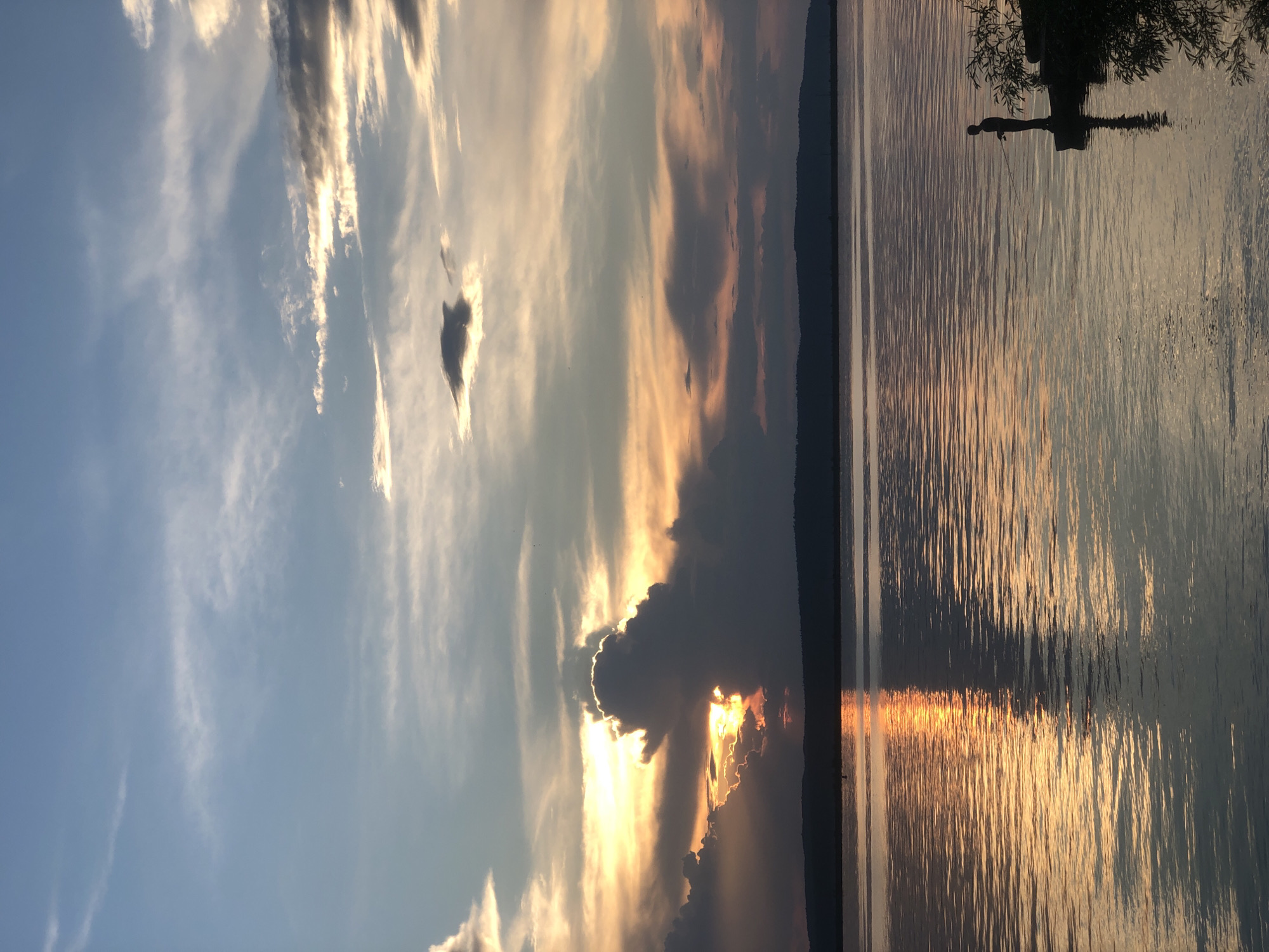 The image depicts a serene sunset over the Kama River in Chaikovsky, Russia. A solitary figure stands on a rocky outcrop, silhouetted against the water as they cast a fishing line. The sky is a canvas of dramatic clouds, with the setting sun peeking through, casting a warm, golden glow that reflects brilliantly on the rippling surface of the river. In the distance, the dark silhouette of a distant shore with rolling hills is visible. The overall atmosphere is one of tranquility and natural beauty.