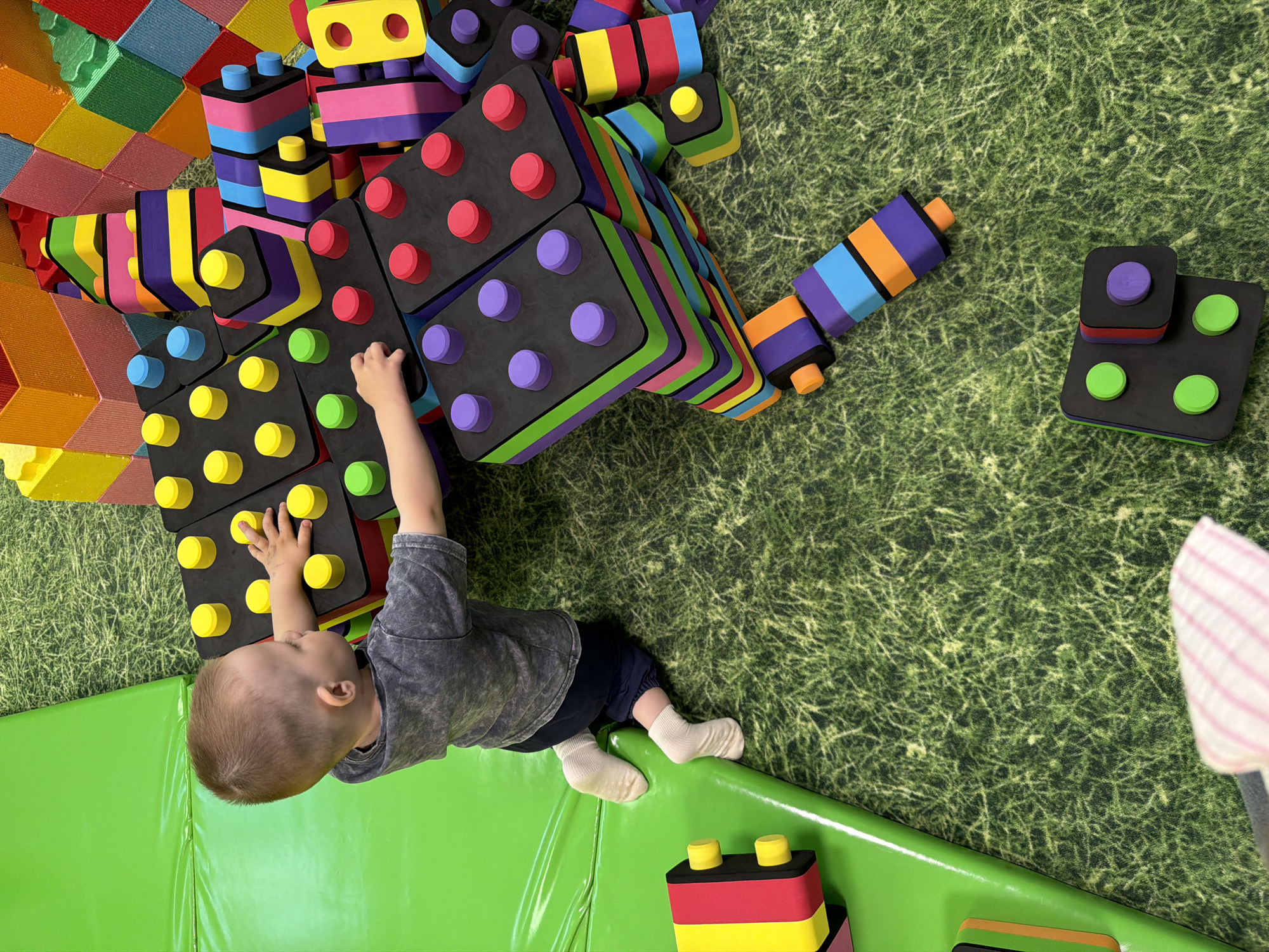 A high-angle shot captures a child playing with large, colorful foam blocks in a room likely in Chaikovsky, Russia. The child, wearing a gray shirt and dark pants, is on their back, reaching towards a tower of black blocks with yellow pegs. The blocks are part of a larger arrangement of various shapes and colors, including stacked, cylindrical blocks in rainbow hues. The floor is covered in a patterned green material resembling grass, with a bright green padded surface bordering the bottom of the frame. In the lower right, part of a light pink object is visible. Another block with yellow pegs sits near the bottom edge of the image.