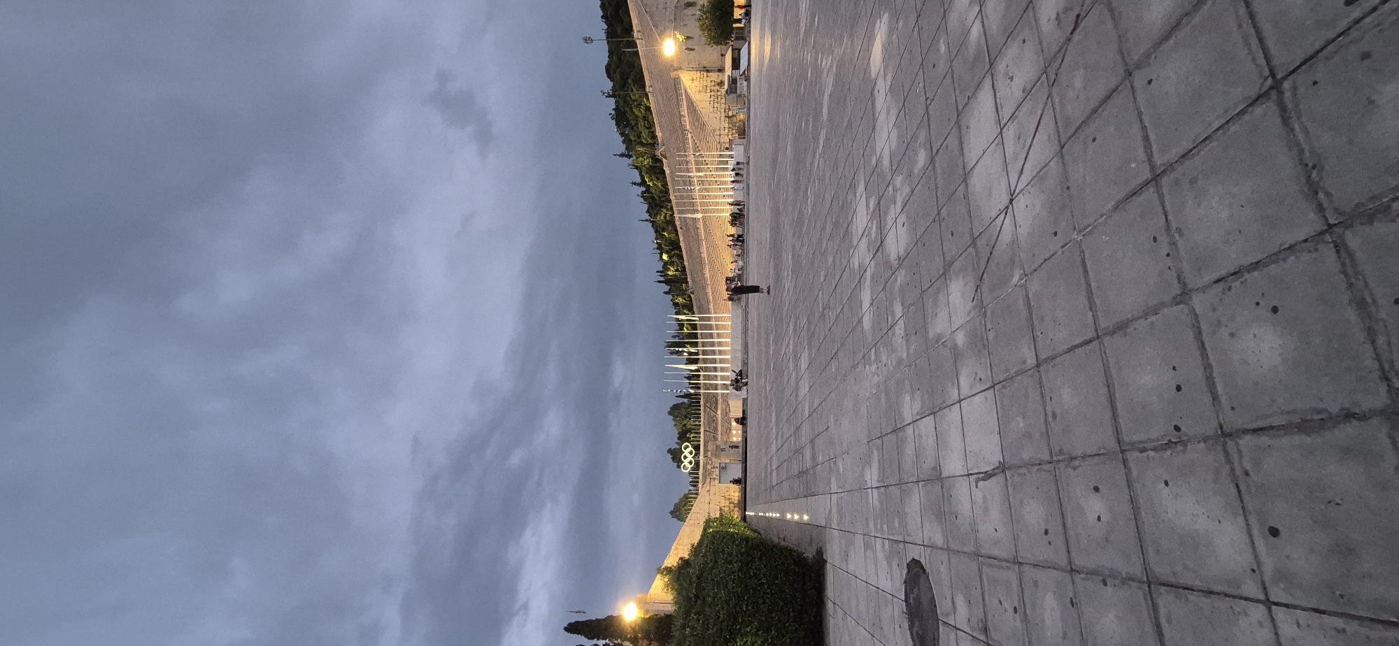 The image depicts the Panathenaic Stadium in Athens, Greece, at dusk. The stadium is characterized by its marble seating, which is illuminated by artificial lights, creating a warm glow against the darkening sky. A prominent display of the Olympic rings is visible atop the stadium's structure. In the foreground, a large, paved plaza extends outward, composed of square, grey concrete tiles. Several individuals are present in the plaza and within the stadium seating area, appearing to be visitors or spectators. The sky is overcast with heavy, grey clouds. Lush green hedges border the left side of the plaza, and trees line the upper slopes of the stadium. The overall atmosphere suggests a twilight scene at a historical landmark.