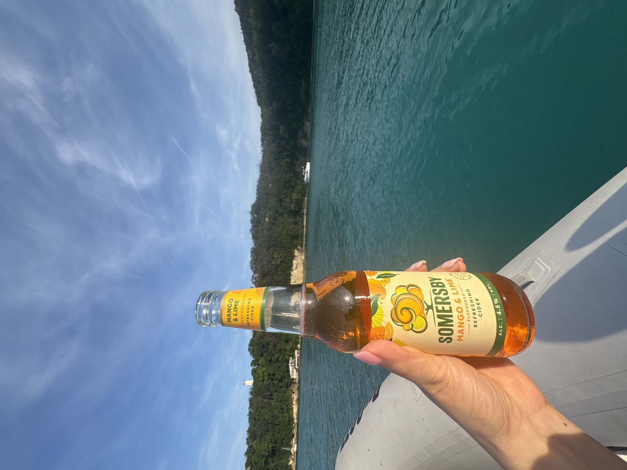 A person's hand, with light skin and light pink manicured fingernails, holds a clear glass bottle of Somersby Mango & Lime Flavoured Refreshing Cider. The bottle's label displays "MANGO & LIME", "FLAVOURED REFRESHING CIDER", and "ALC. 4.5% VOL.". The cider within the bottle is an amber-orange liquid. The hand and bottle are positioned over the white inflatable tube of a boat or dinghy, which occupies the lower left quadrant of the image.
The boat is situated on a body of calm, turquoise-green water exhibiting subtle ripples. In the background, a heavily forested coastline extends across the horizon. On the left side of the coastline, a white lighthouse-like structure is visible on a hilltop, with a smaller white building featuring a red roof nestled among the trees below it. A white sailboat is moored or anchored in the water near this section of the shore. To the right, further out on the water, a white powerboat or yacht is present against the dense green foliage of the distant shore. The sky above is a clear blue with scattered thin white clouds. The scene is illuminated by bright daylight, casting visible shadows. The location is in the city of Unknown, Unknown.