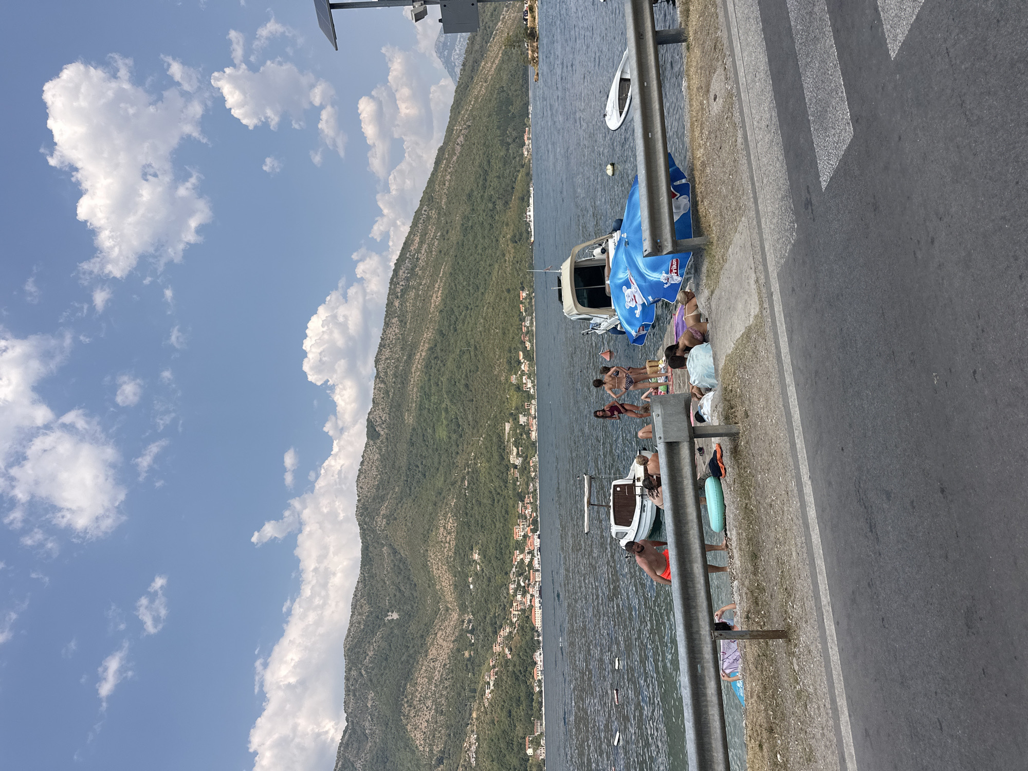 The image captures a sunny day at the waterfront in Lepetane, Montenegro. A calm body of water, likely a bay or a section of the sea, is visible, with several boats docked or floating near the shore. The shore itself is a mix of gravel and concrete, with a metal guardrail separating it from a paved road in the foreground.
Several people are seen enjoying the water and the sun. Some are standing in the shallow water, while others are relaxing on the shore. A few individuals are lying on towels or inflatable mats, with one person taking a photo. There are at least three boats in the water: a small white boat with a canopy, a larger blue inflatable float with a cartoon image, and a smaller white boat positioned further out.
In the background, a lush green mountain slopes down towards the water, dotted with red-roofed buildings, suggesting a residential area. The sky is a clear blue with scattered white clouds, indicating fair weather. The overall scene depicts a typical summer day at a coastal recreational area.