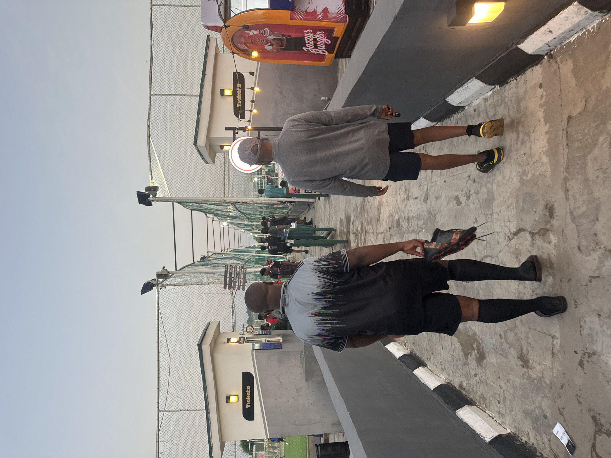 The image depicts two men standing on a paved area, likely an outdoor entertainment or sports facility, in Abuja, Nigeria. The overall orientation of the image is sideways, with the subjects appearing to be standing upright relative to each other.
The foreground subject is a man wearing a black and grey t-shirt with a graphic design, black shorts, and black socks with black shoes. He is holding a black and orange athletic shoe in his right hand. The second subject, slightly further back, is wearing a grey long-sleeved shirt, dark shorts, black socks, and black and yellow sneakers. He is also wearing a light-colored baseball cap.
The setting appears to be an area with a chain-link fence in the background, possibly a driving range or sports court. There are structures with lighting, including a sign that reads "Tickets". Further back, another sign is visible with "Tickets" written vertically and illuminated lights. There's also a colorful advertising structure with "Jazzy's Burger" printed on it. Other people are visible in the background, suggesting a public space. The sky is a pale blue, indicating it could be daytime. The ground is concrete with some wear and tear, and there is a black and white striped curb. The atmosphere seems relaxed and casual, possibly during a leisure activity.
