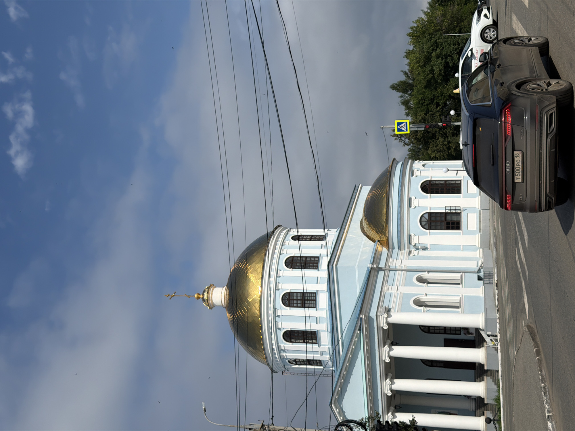The image shows a light blue church with a golden dome and a cross on top, located in Votkinsk, Russia. The church has white columns and arched windows. In front of the church, there is a street with two cars, a dark gray SUV and a white car. There is also a pedestrian crossing and a traffic light. The sky is blue with some clouds.