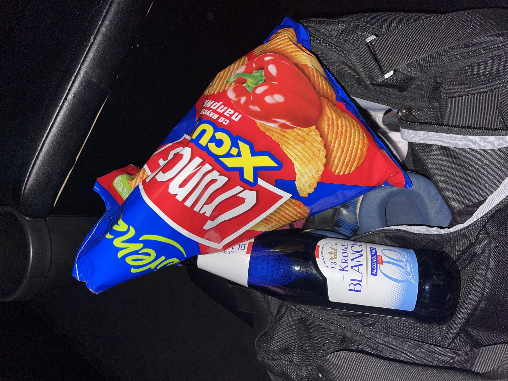 The image displays a snack and beverage setup within a dark car interior. A prominent, vibrant red and blue bag of chips rests partly on a black duffel bag. The chip bag features a large image of a red bell pepper, and visible text includes "Crunc XCU," "со вкусом паприки" (Russian for "with taste of paprika"), and partially "rena."
Next to the chip bag, inside the open black duffel bag, lies a dark blue bottle. Its white and blue label clearly shows "KRONE BLANCHE," "ALCOHOL FREE," and "BIERE." Additional text on the bottle reads "WITH A HINT OF CITRUS" and includes a crown logo along with the numbers "13 | 18." The duffel bag itself is made of a textured black material, featuring zippers and straps.
The surrounding environment is dark, suggesting an evening or nighttime setting, or an interior with limited light. Parts of the car's interior, including a black leather-like surface with a seam and a circular cup holder, are visible on the left. The scene implies a casual stop for refreshments, possibly while traveling or after an activity, located in Khimki, Russia.