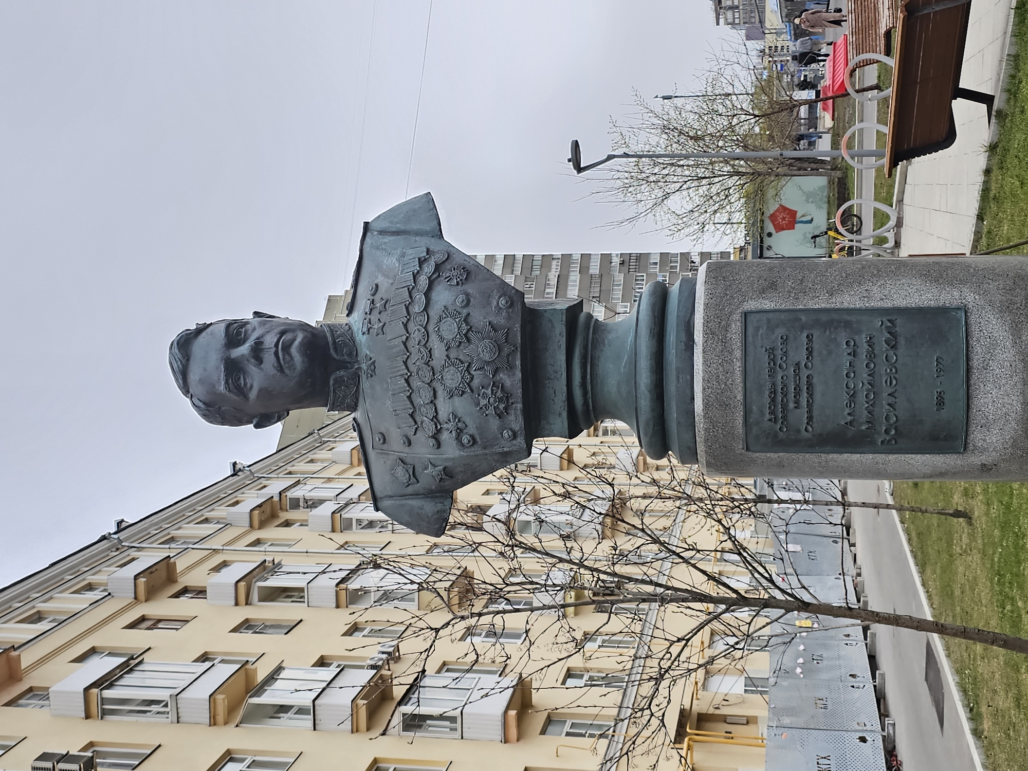 A bronze bust monument of a military figure is the central subject of this image, positioned outdoors on a grey, overcast day. The bust depicts a stern-faced man in uniform, heavily adorned with numerous medals and orders on his chest and epaulets. It is mounted on a rectangular grey stone pedestal.
A dark metal plaque is affixed to the front of the pedestal, displaying Cyrillic text. The inscription reads: "Дважды Герой Советского Союза" (Twice Hero of the Soviet Union), "Маршал Советского Союза" (Marshal of the Soviet Union), "АЛЕКСАНДР МИХАЙЛОВИЧ ВАСИЛЕВСКИЙ" (Aleksandr Mikhailovich Vasilevsky), followed by his birth and death years: "1895 - 1977".
In the background to the left of the monument, a multi-story building with a light yellow facade and numerous windows is visible, many of which have white, box-like enclosures or structures beneath them. A bare-branched tree stands in front of the building, partially obscuring it. To the right of the bust, a public pathway runs alongside a strip of green grass. A wooden bench with red side supports and several white, circular bicycle racks are present along the path. People are seen walking in the distance. A light pole with a circular fixture and another bare tree are also visible. Further back, a row of grey, slatted utility boxes or fencing is present, with the letters "КТХ" legible on some panels. The scene depicts a static memorial in an urban park or square setting under a light grey sky.