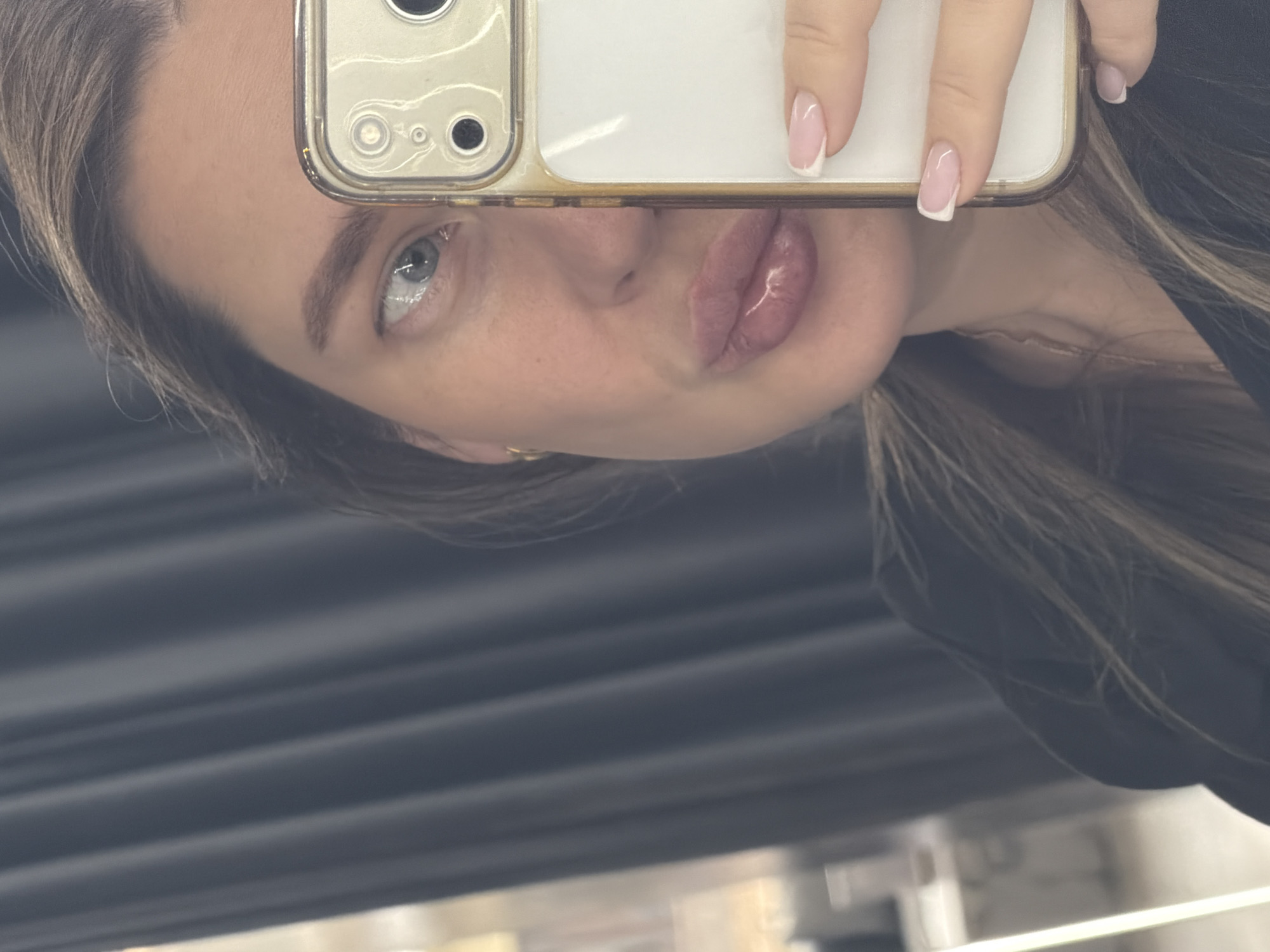 A woman holds a smartphone up in a selfie style, partially obscuring her face. She has fair skin, light eyes, and is wearing a black top. Her nails are manicured with a French tip design. The background is blurry, suggesting an indoor setting with horizontal lines, possibly window blinds or a textured wall. The location is not identifiable as Moscow, Russia based on visual information.