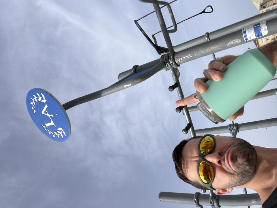 A low-angle shot features a man with sunglasses holding a green water bottle next to a metal structure. His finger points toward the structure. Above, a blue sign with a white laurel wreath and the letter "V" is attached to a pole. The sky is visible in the background, with some clouds. The reflection in the man's sunglasses shows a yellow building. The perspective gives a distorted view of the scene.