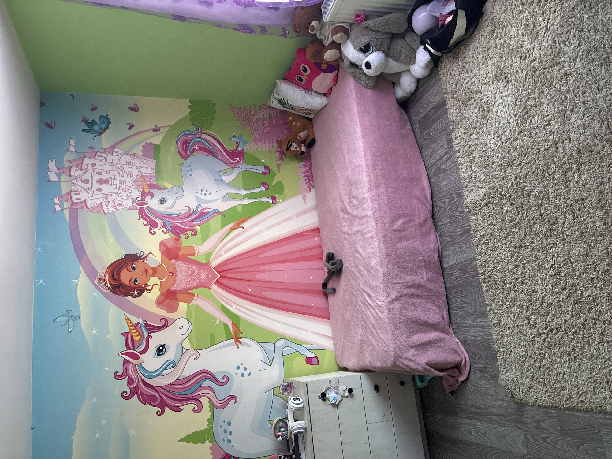 A child's bedroom is depicted. The walls are painted with a mural featuring a princess, unicorns, and a castle. A pink-covered bed is in the center of the frame with several stuffed animals arranged on the head. A rug lies on the floor. A white nightstand sits next to the bed. The location is Aldomirovtsi, Bulgaria. There are no people in the image.
