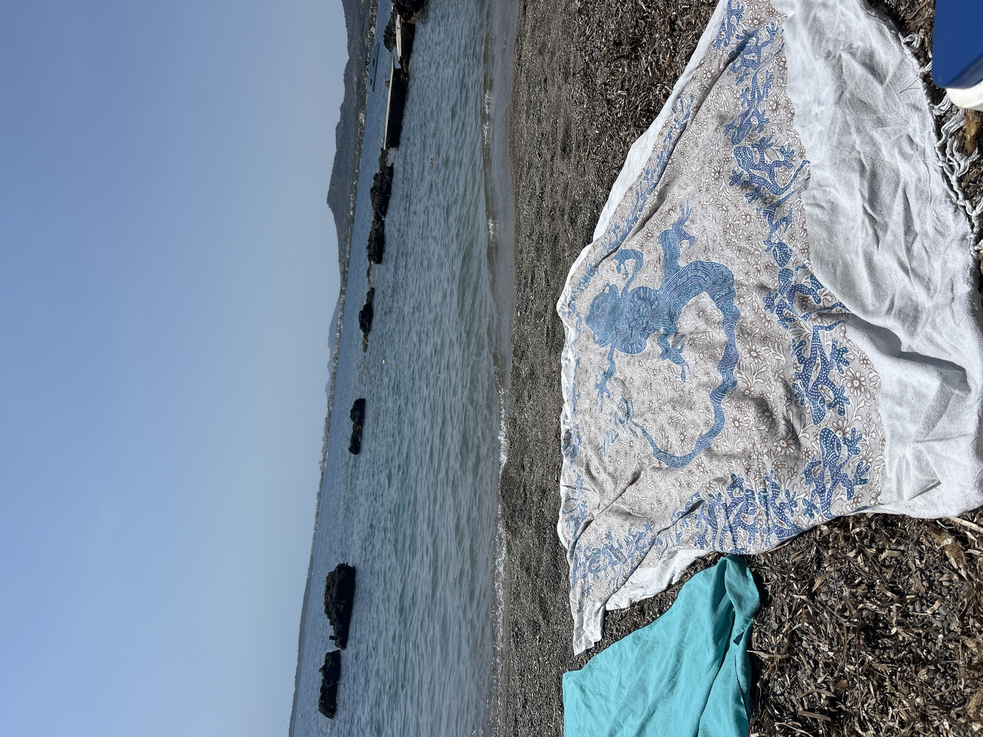 The image captures a scene on a beach in Costa D'en Blanes, Spain, under a clear blue sky. In the foreground, two beach towels are laid out on dark, pebbly sand mixed with dried seaweed. The towel on the left is a solid turquoise color. The towel on the right is white with a decorative pattern in shades of blue and brown, featuring stylized figures that appear to be lizards or similar creatures.
Beyond the beach, the calm, blue sea stretches towards a distant shoreline. Several dark, irregular-shaped rocks are partially submerged in the water. In the middle ground, a small wooden pier or jetty extends into the sea. The background shows a town or village nestled along a hilly coastline, with buildings visible. A white yacht is moored in the water to the right of the pier. The overall impression is a peaceful, sunny day at the beach.