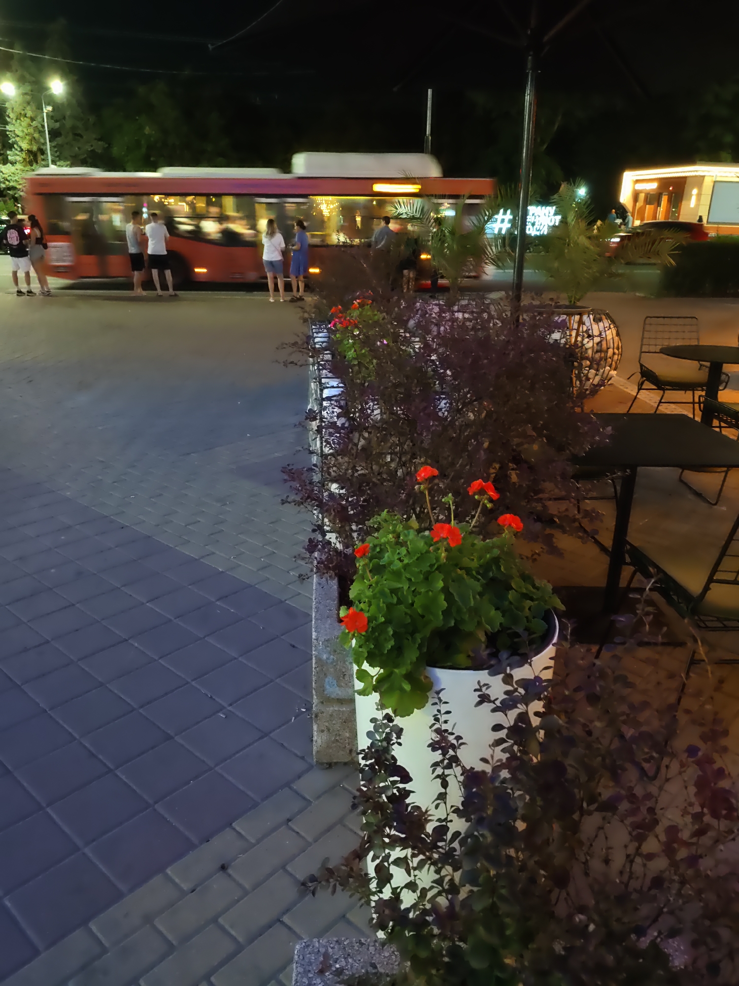 The image depicts a nighttime scene in an urban setting, likely outdoors near a sidewalk cafe or waiting area. A red bus is in motion on the left side of the frame, with blurred lights indicating its movement. Several people are standing near the bus, appearing to be waiting or disembarking. In the foreground, there are planters filled with green foliage and red flowers. To the right, outdoor seating with tables and chairs is visible, suggesting a commercial establishment. The ground is paved with interlocking stones. The overall atmosphere is dimly lit, with artificial lighting from streetlights, the bus, and the cafe area.