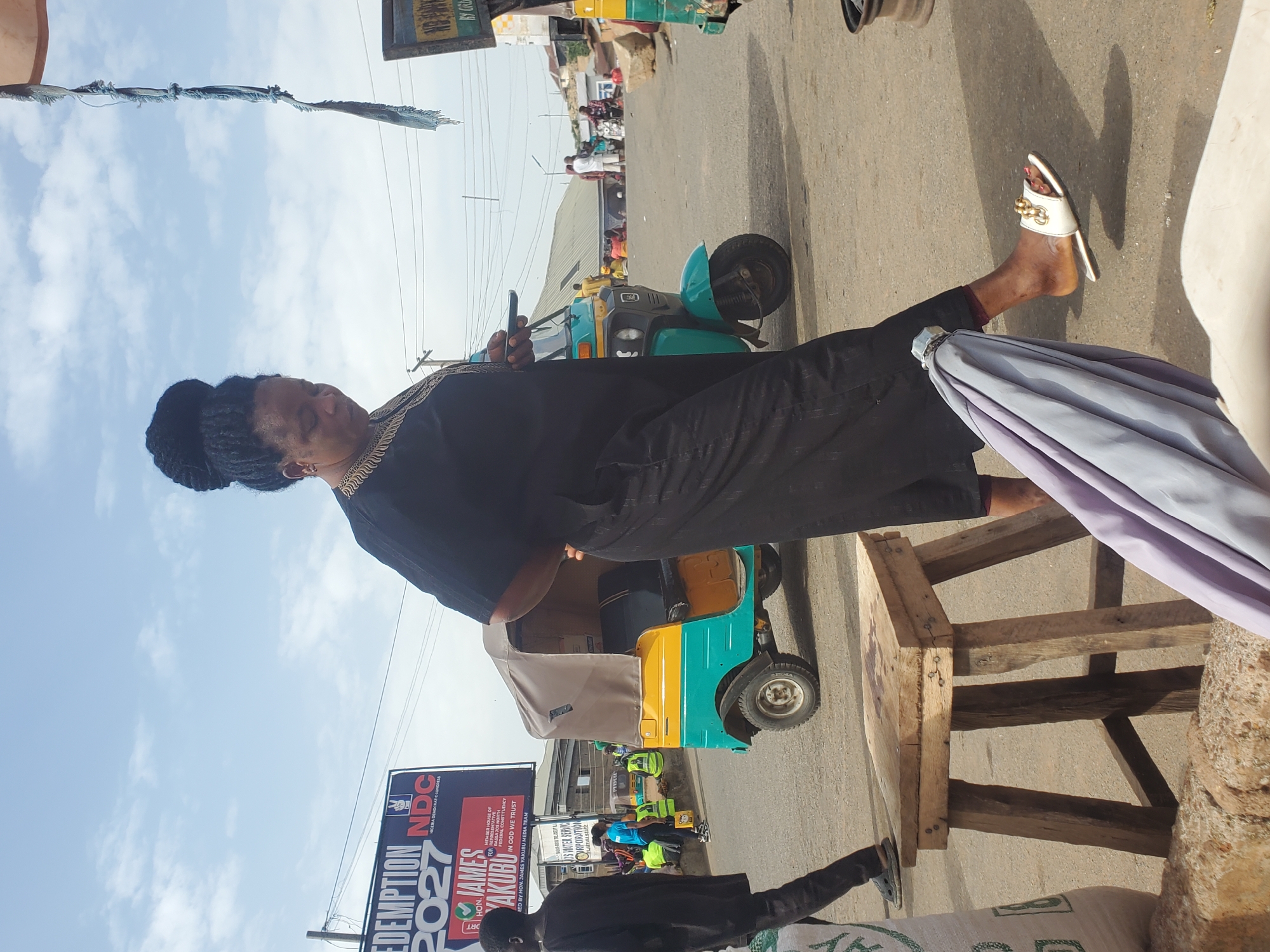 In the city of Gwong, Nigeria, a woman with her hair in a bun, wearing a black dress with gold trim, stands looking at her phone. She is wearing white sandals with gold accents and is holding a purple umbrella. Next to her is a wooden stool. In the background, a yellow and teal auto-rickshaw is parked on a paved road. Several people are visible in the distance, including a young man in a black outfit walking to the left and others near a billboard. The billboard, which has "REDEMPTION 2027" and "NDC NIGERIA DEMOCRATIC CONGRESS" written on it, is a prominent feature of the scene. The sky is cloudy.