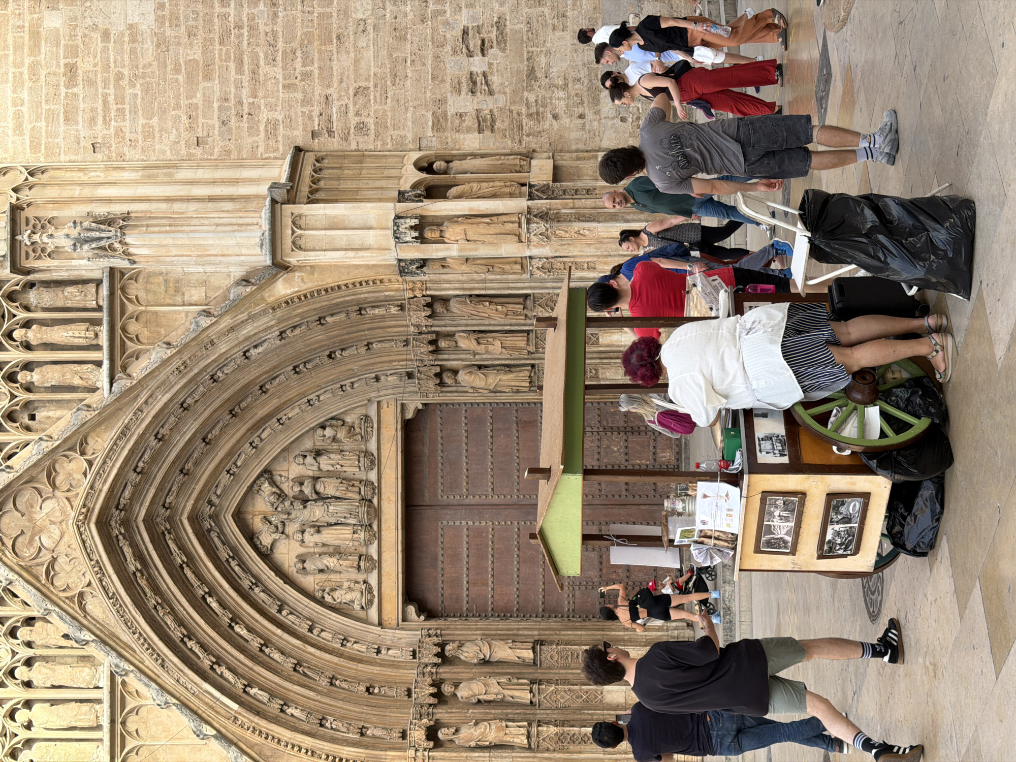 The image depicts an outdoor scene in front of a large, ornate stone building, possibly a cathedral, with intricate architectural details and sculptures. In the foreground, a mobile food cart with a green roof is set up, attended by several people. A woman in a white shirt and striped skirt is interacting with the cart. Various individuals are standing around the cart, some appearing to be customers, while others are walking past. To the left, two men are standing near the entrance of the building. The ground is paved with cobblestones. The overall atmosphere suggests a bustling public space.