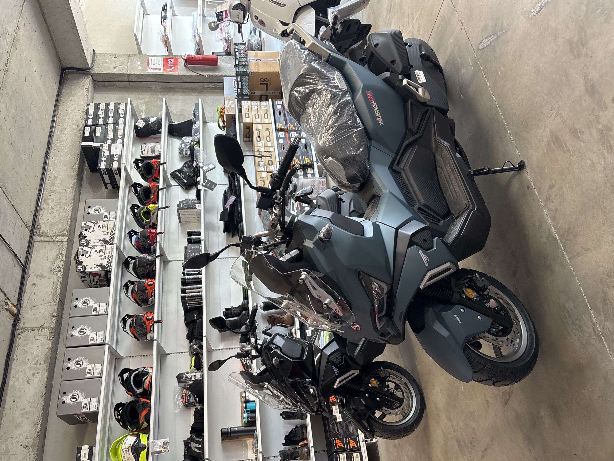 In a store in Tbilisi, Georgia, two scooters are displayed on a concrete floor. The scooter in the foreground is dark teal and has a plastic cover over its seat. The scooter behind it is black. Shelves line the wall behind the scooters, displaying various motorcycle helmets and other gear. A fire extinguisher is visible on the right side of the frame.