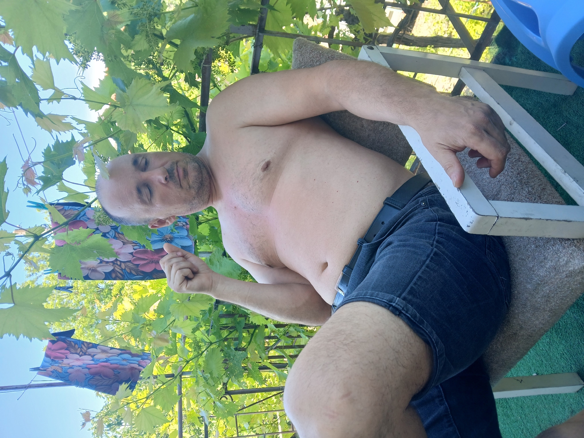 The image depicts a bare-chested man with short hair, wearing dark denim shorts and a black belt, sitting on a chair. He is outdoors, with lush green foliage and a bright blue sky visible in the background. Two pieces of fabric with floral patterns are hanging on a clothesline to his left. The scene appears to be in a residential yard, potentially in the city of Obrochishte, Bulgaria, given the style of the chair and the surrounding greenery. The man is looking downwards with his eyes closed, and his right hand is raised slightly near his head. A blue plastic object is partially visible in the lower right corner of the frame.