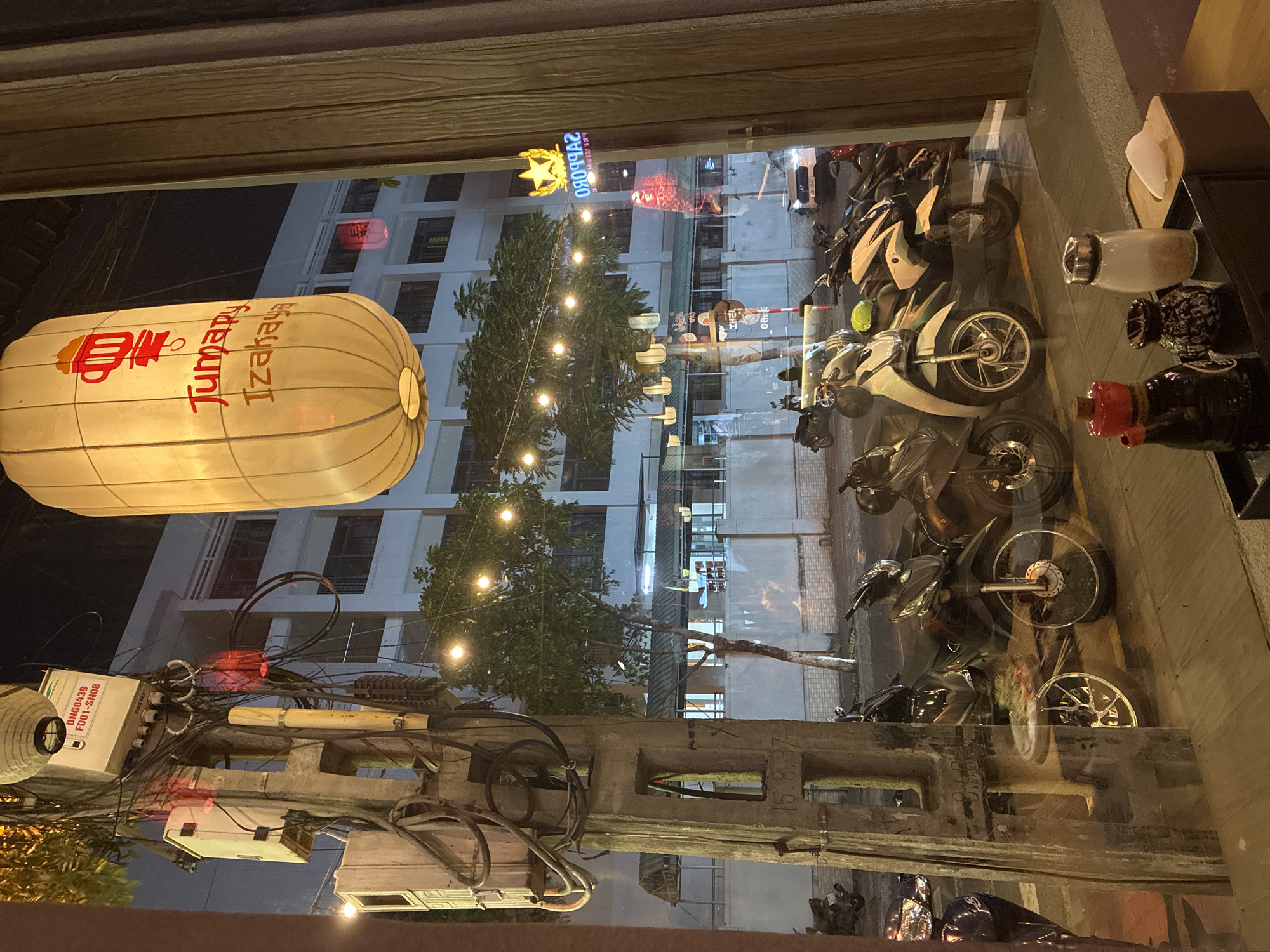 A nighttime view from inside a building looking out onto a street in Đà Nẵng, Vietnam. The view is through a window, with reflections of objects inside the building visible on the glass surface. Outside, several motorcycles are parked along the street. A Japanese lantern with "Jumapy Izahaya" in red lettering hangs from a utility pole cluttered with electrical wires. A building with lit windows rises in the background, and a lighted tree with small bulbs adds to the ambiance. A Sapporo beer sign and a koi-fish kite also hang above the street. A concrete utility pole with visible date markings supports the tangled wires. The scene captures a slice of urban life in Đà Nẵng.