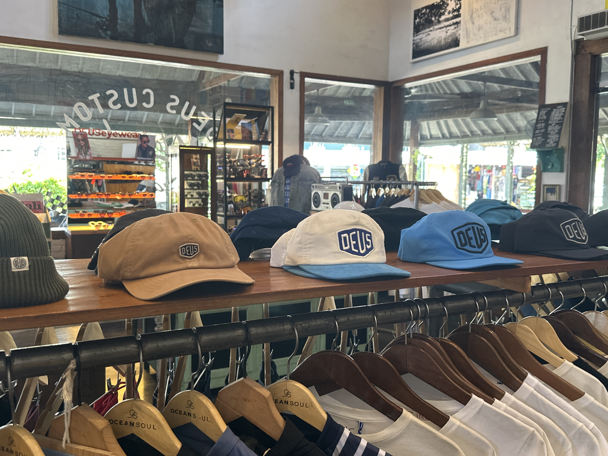 The image captures a retail setting in North Kuta, Indonesia, showcasing apparel and accessories. Prominently displayed on a wooden shelf are several baseball caps and a beanie, all bearing the "DEUS" logo. Below the shelf, a rack holds an assortment of shirts hanging on wooden hangers, some with the label "OCEAN SOUL" visible. In the background, a storefront with reversed text "USeyewear" is visible through the windows. A boombox and various clothing items are also present, adding to the store's ambiance.
