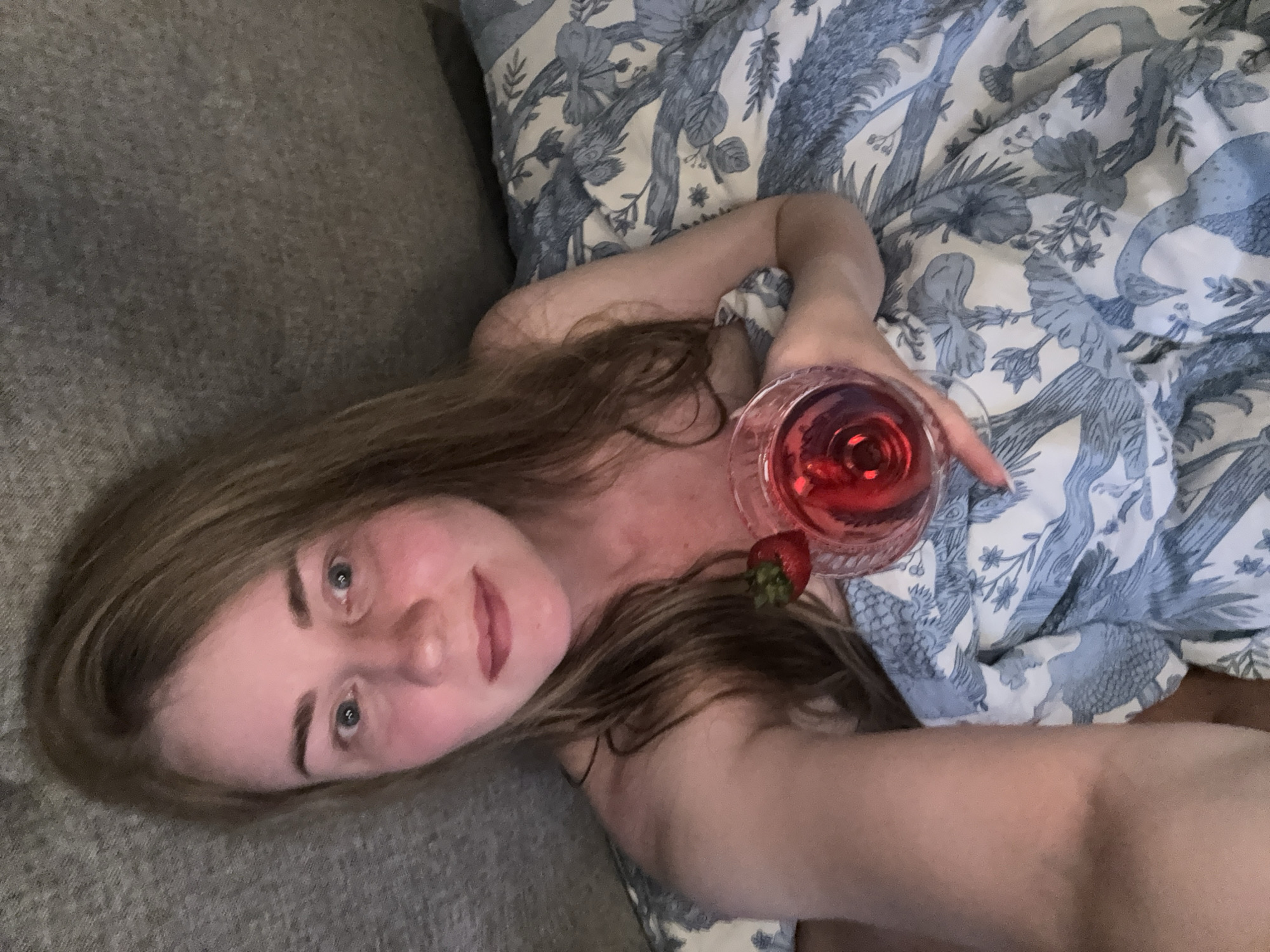 A light-skinned female lies on her left side, partially covered by a patterned blue and white duvet, next to a gray couch. She holds a stemmed glass filled with red liquid in her right hand, accompanied by a strawberry. The location is indeterminable from the image and cannot be placed in Saint Petersburg, Russia.