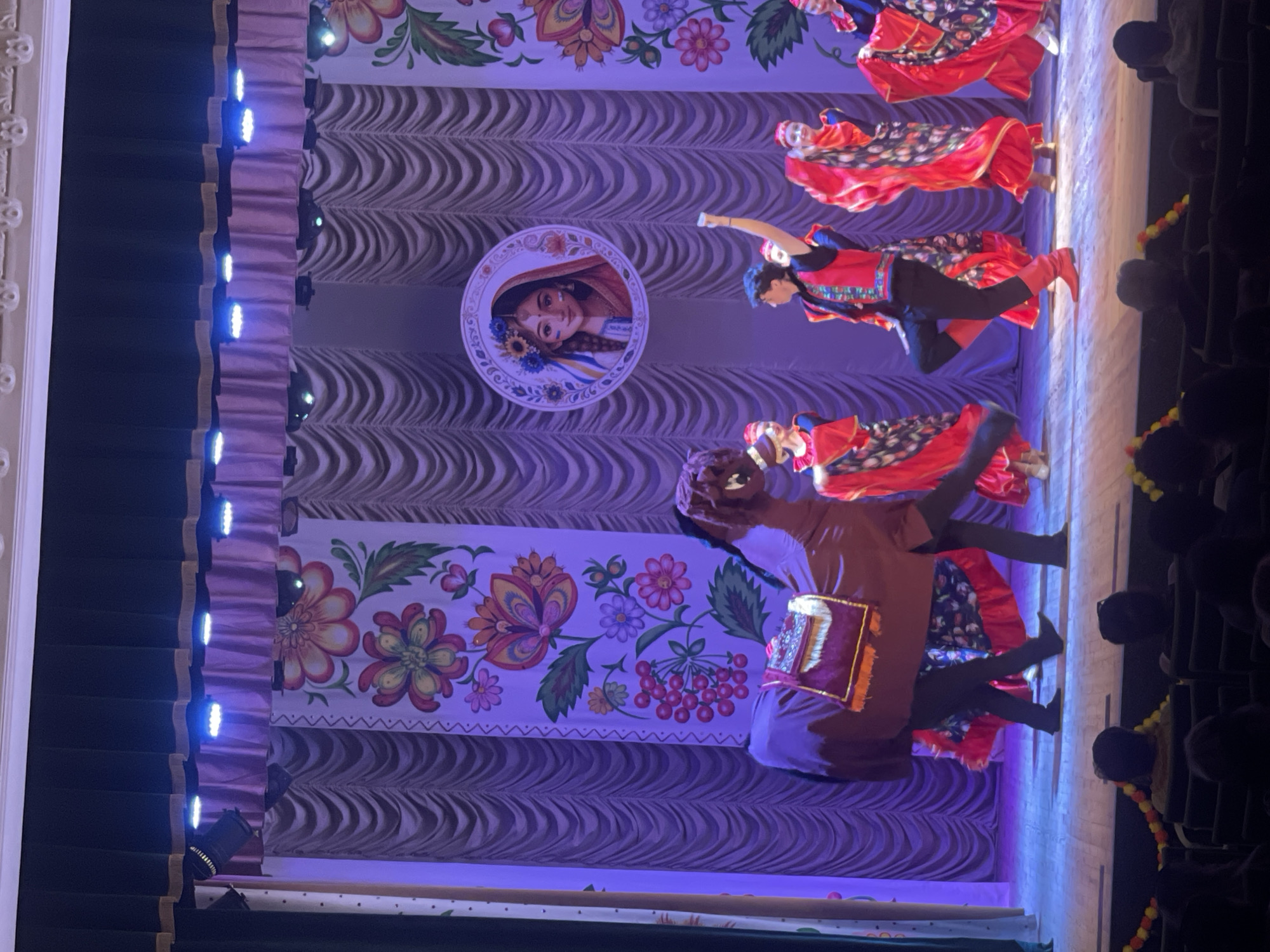 A performance is taking place on a stage in Гожули, Ukraine. The stage is adorned with floral-patterned curtains and a circular graphic of a woman with flowers in her hair. Several performers, dressed in traditional red and black folk costumes, are dancing. One performer is dressed as a horse, with two people inside. The audience is visible in the foreground, seated in a dark auditorium. The lighting is primarily blue and purple, with some white spotlights.