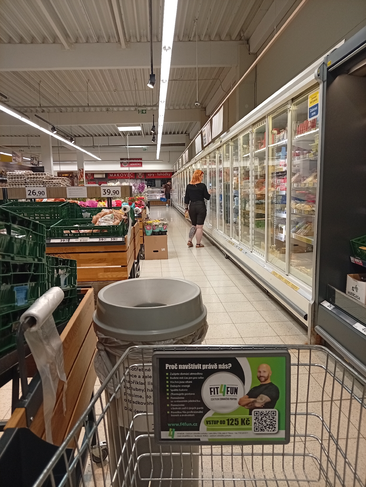 This image depicts the interior of a supermarket in Tišnov, Czechia, with a focus on the checkout area and a promotional display attached to a shopping cart. The lighting is bright, typical of a retail environment, with long fluorescent tubes running along the ceiling. In the foreground, a shopping cart is visible, and attached to its handle is a digital advertisement featuring a bald man with tattoos, a logo that reads "FIT 4 FUN CENTRUM ZDRAVÉHO POHYBU", and a QR code. The advertisement also lists health-related benefits and a starting price of "VSTUP OD 125 Kč".
To the left of the cart, a grey trash bin with a trash bag lining is present. Further back, rows of grocery shelves and refrigerated display cases extend into the distance. Several rows of green plastic crates, some filled with produce or other goods, are stacked in front of wooden shelving units. In the mid-ground, a woman with orange hair is seen browsing the contents of a refrigerated display case. Various price tags and signs are visible throughout the store, indicating products and promotions. The floor is tiled with a light-colored pattern. The overall scene suggests a typical shopping experience in a modern supermarket.