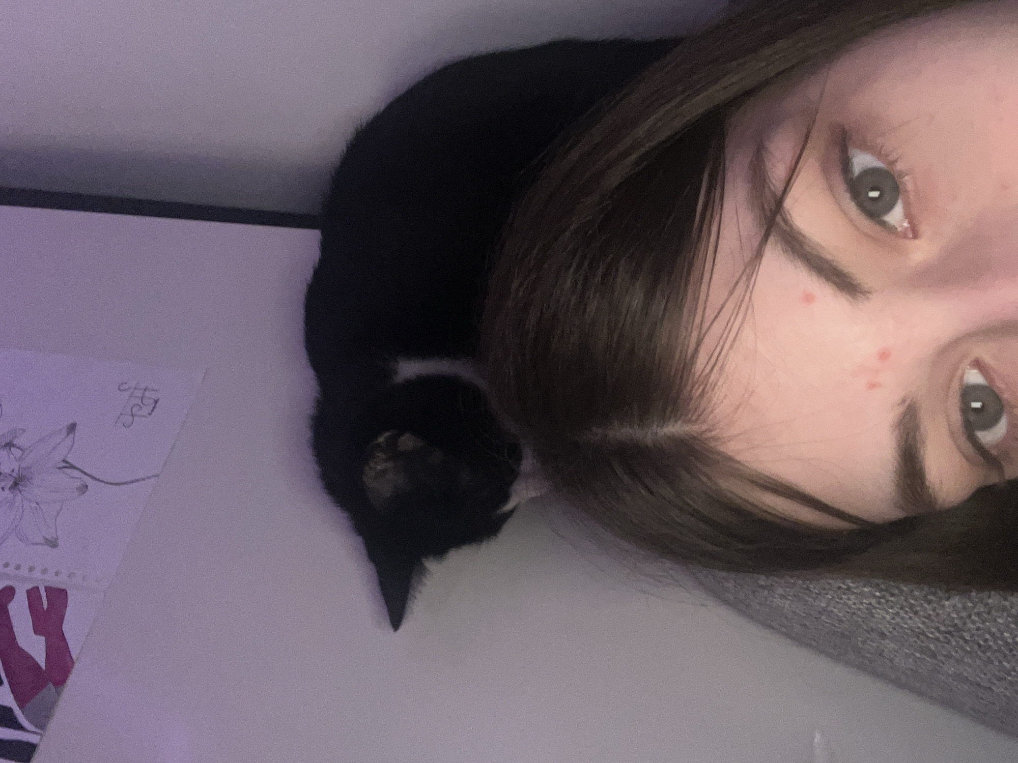 A close-up, slightly angled shot shows a woman with fair skin and dark brown hair. Her face is turned to the right, with one eye visible and a portion of her forehead, eyebrows, and nose in view. Several small red spots are visible on her face. Next to her head is a black cat. The cat's tail and body are positioned towards the left side of the image. The background includes a surface which appears to have a drawing of a flower and other indistinct shapes. The environment appears to be indoors. There is no verifiable information indicating the location as Saint Petersburg, Russia.