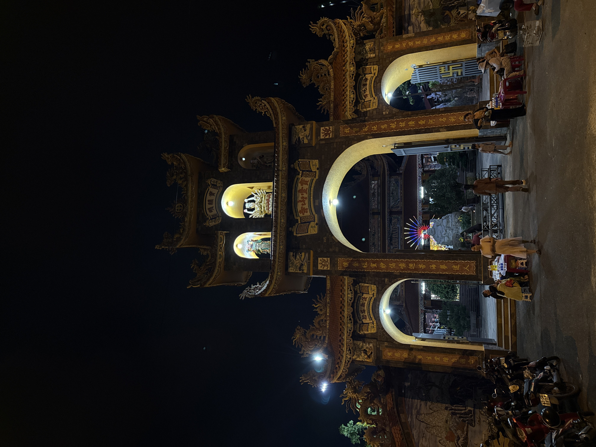 At night, a low-angle, sideways shot shows the entrance to a Vietnamese temple, likely in Đà Nẵng. The entrance structure is adorned with golden statues of dragons. People walk and stand in the gateway's archway. Motorbikes are parked at the base. Lanterns and carvings adorn the structure. Inscriptions in Chinese and Vietnamese are visible on the entrance walls.