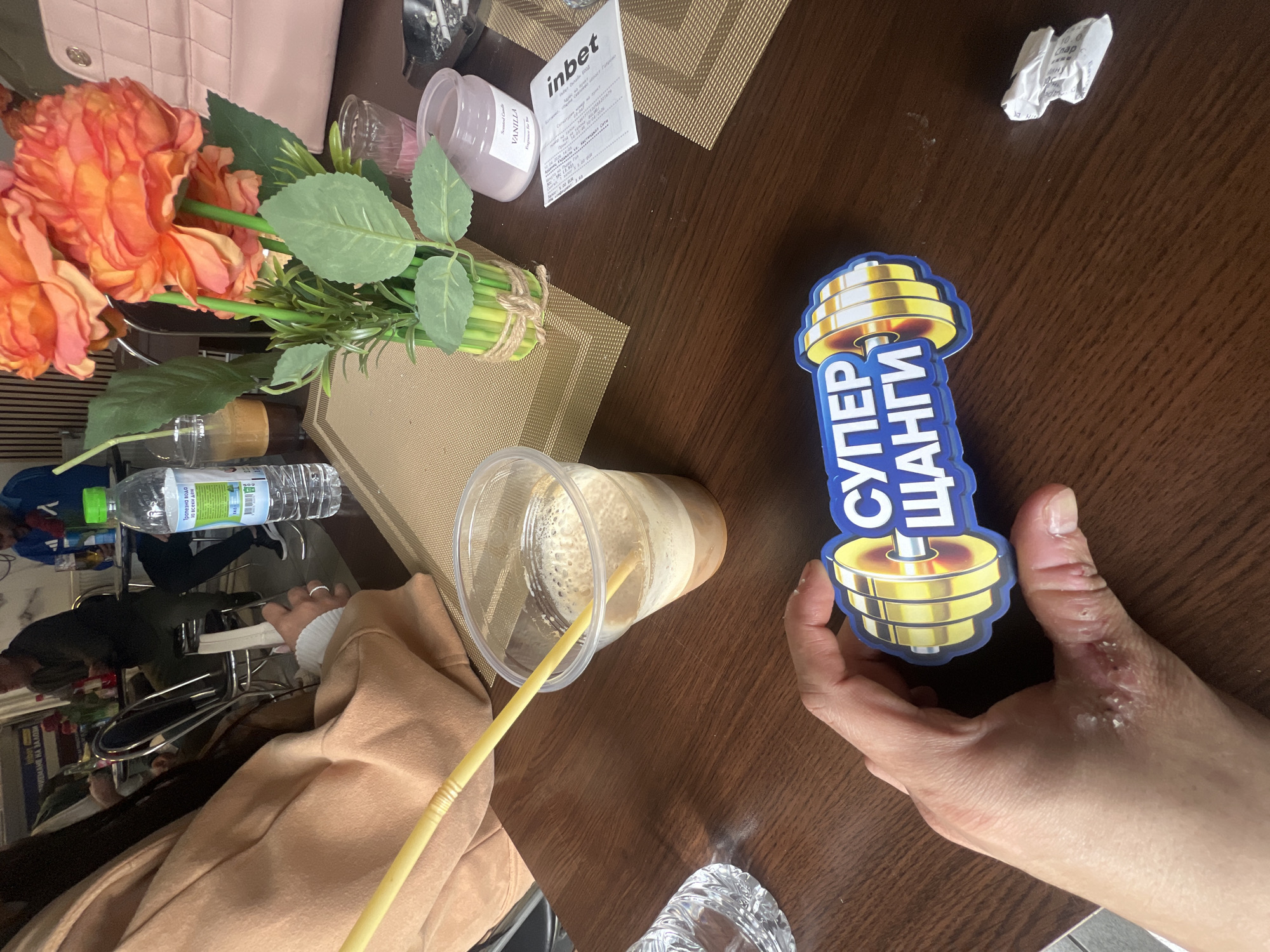 The image shows a table setting in what appears to be a cafe or restaurant in Batoshevo, Bulgaria, featuring a variety of objects, including artificial flowers, a beverage in a clear plastic cup with a yellow straw, a bottle of water, a Vanilla-scented candle, a betting slip from "inbet", and a sticker with Cyrillic text and an image resembling a dumbbell. A person's hand is holding the sticker, suggesting an interaction with the objects on the table. The setting appears to be indoors, with elements of daily life and leisure activities present.