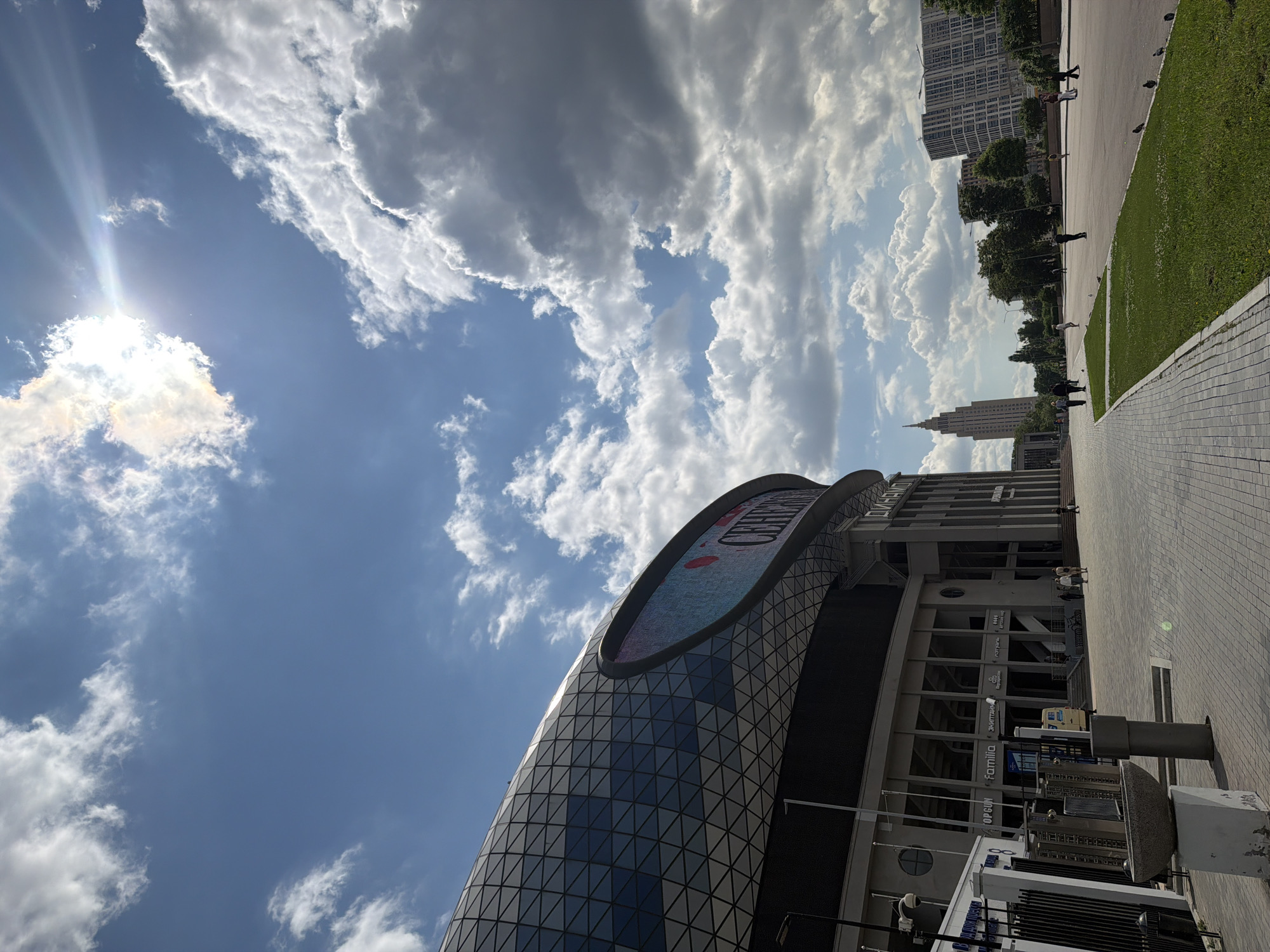 A low-angle, full shot captures the exterior of a large, modern building with a curved, faceted facade and a large LED screen. The building appears to be a sports arena or stadium, with the word "ДИНАМО" visible on the screen. The facade is made of glass panels arranged in a triangular pattern, reflecting the bright blue sky and scattered white clouds. The sun is prominently positioned in the upper right corner, casting rays through a gap in the clouds.
In the foreground, a paved walkway leads towards the building, bordered by a strip of green grass with several pigeons foraging. Several people are walking along the path and in the distance. To the left of the walkway, a security turnstile system is visible, along with a concrete planter and a light pole.
In the background, a tall, slender skyscraper with a spire stands against the skyline, along with other mid-rise buildings. The overall environment suggests an urban setting, possibly a park or plaza adjacent to the stadium, under a partly cloudy sky. The lighting indicates it is daytime, likely mid-morning or afternoon.