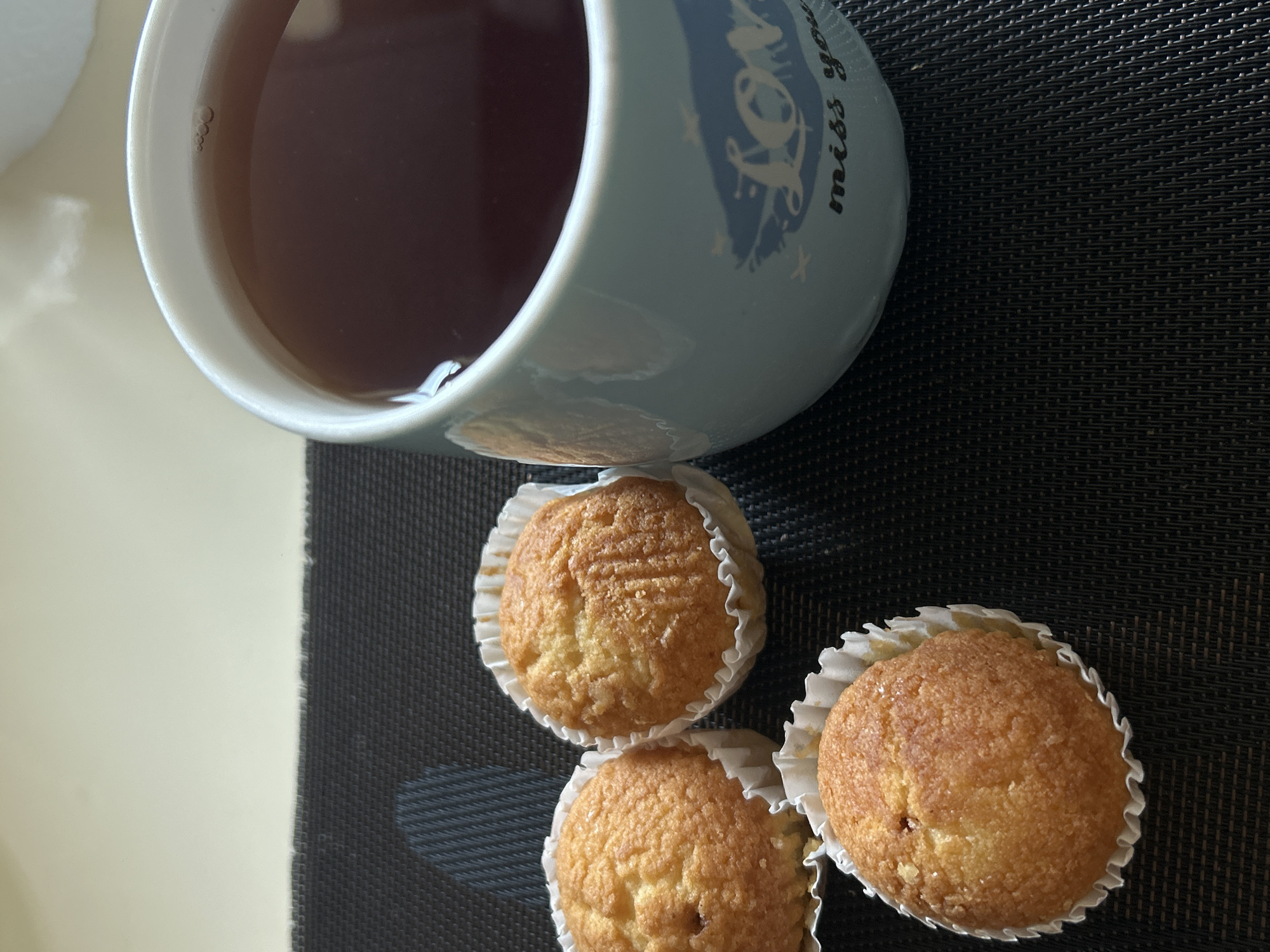 A close-up, eye-level shot displays a still life of baked goods and a beverage. In the foreground, three golden-brown muffins, nestled in white paper liners, are arranged on a dark, textured placemat. The muffins have a slightly domed top with a subtly textured surface. To the right, a light blue mug filled with dark amber liquid, likely tea, occupies the majority of the frame. The mug has a visible design with the word "Love" and the phrase "miss you" printed in a dark color. The background consists of a pale, textured surface, possibly a table, with a thin, horizontal line separating it from the placemat. The lighting suggests an indoor setting, with soft illumination highlighting the objects. The scene is devoid of human presence and appears to be located in Khyrdalan, Azerbaijan.