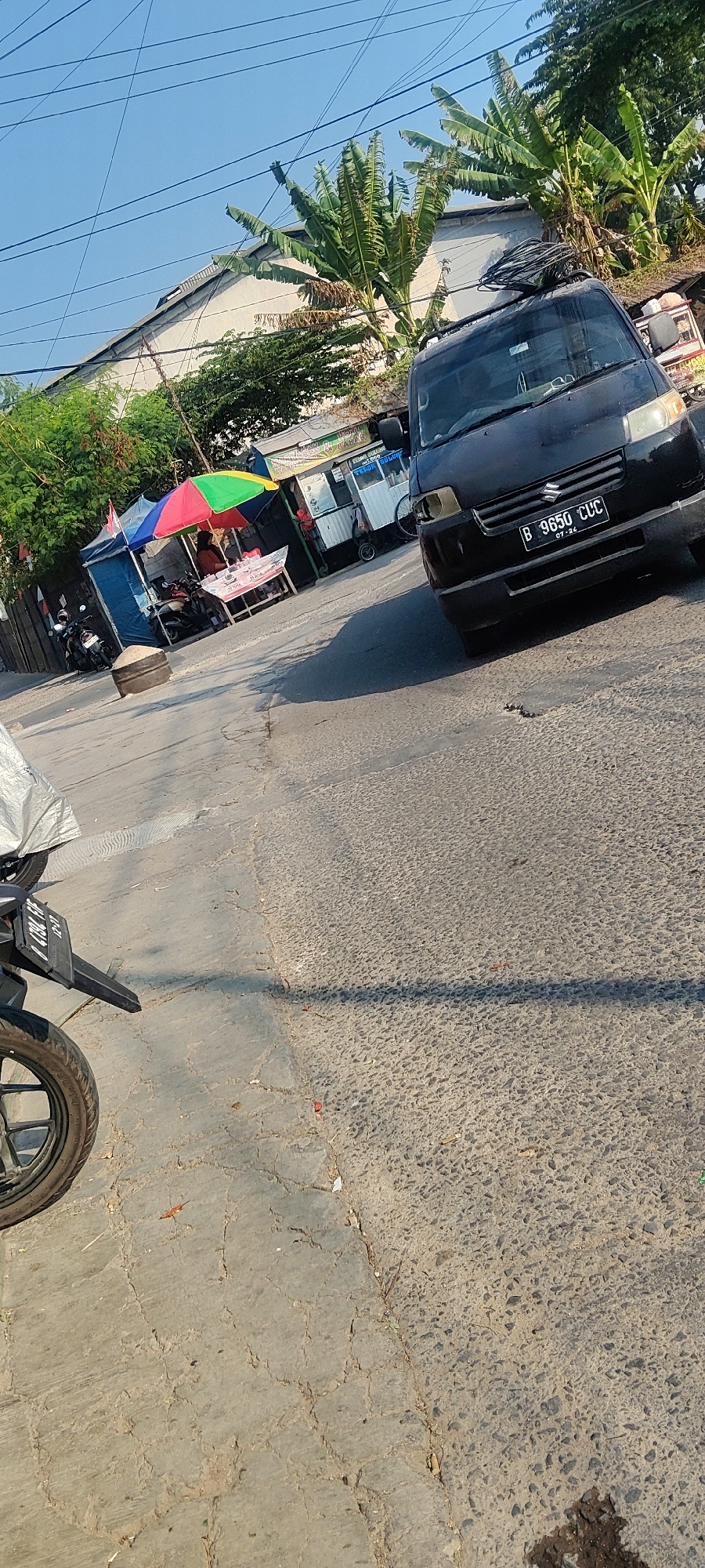 A black Suzuki APV van with license plate B 8650 CUC is parked on a cracked asphalt road in Mekarmukti, Indonesia. The van is facing right, with its headlights on. In the background, there are several food stalls with colorful umbrellas and awnings. A woman is visible behind one of the stalls, tending to customers. Banana trees and other greenery line the street, and power lines are strung overhead. A motorcycle is parked to the left of the stalls. The sky is clear and blue, and the sun is casting shadows on the road.