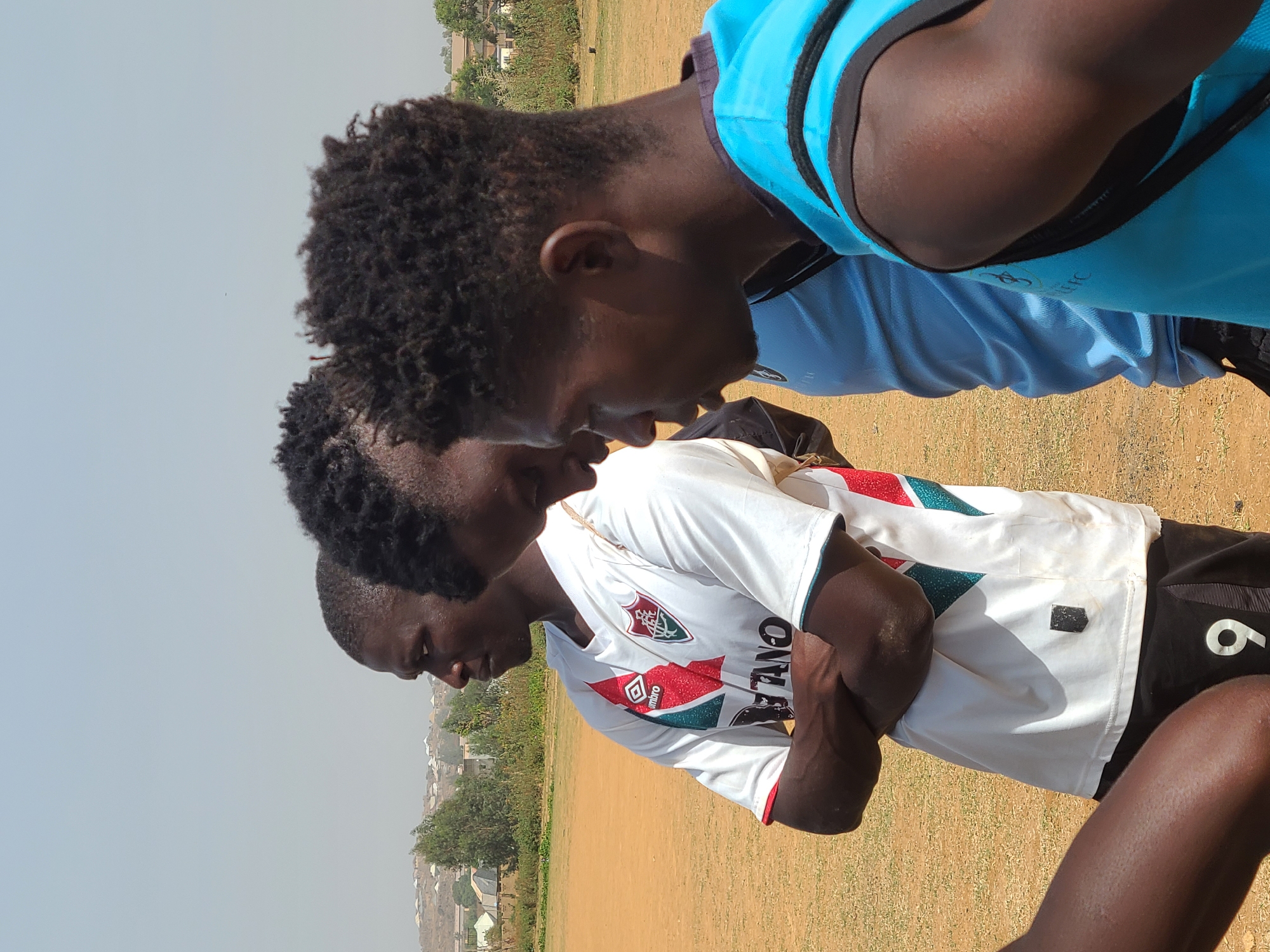 The image, taken in Barikin Jirga Summa, Nigeria, depicts three young men gathered closely on a dry, open field under a bright, hazy sky. The photo appears to be taken outdoors, likely during midday or early afternoon, given the clear sky and lack of strong shadows.
The three subjects are young men of African descent, with dark skin tones and short, curly hair. They are positioned in a close huddle, looking downwards, suggesting they are either listening intently, observing something on the ground, or engaged in a serious discussion. Their expressions appear focused and contemplative.
The man on the left wears a white short-sleeved t-shirt with a distinctive pattern featuring green, red, and white elements. A football club crest is visible, along with the "UMBRO" logo and partially visible text that appears to spell "FLUMINO," likely referring to the Brazilian club Fluminense FC. He is also wearing black shorts with a white number "6" on the thigh. The man in the middle is dressed in a light blue short-sleeved t-shirt with a darker collar and trim, and a subtle logo on the chest. The third man, partially visible on the right, wears a bright blue short-sleeved top with black accents.
The setting is an expansive, unkempt field with dry, brownish-yellow earth, indicative of sparse vegetation or a dirt pitch, common for recreational areas. In the hazy background, distant residential buildings and scattered trees are visible, suggesting a semi-urban or rural environment. The overall scene conveys a casual gathering or a break during an outdoor activity, possibly football, given the athletic attire worn by the individuals.